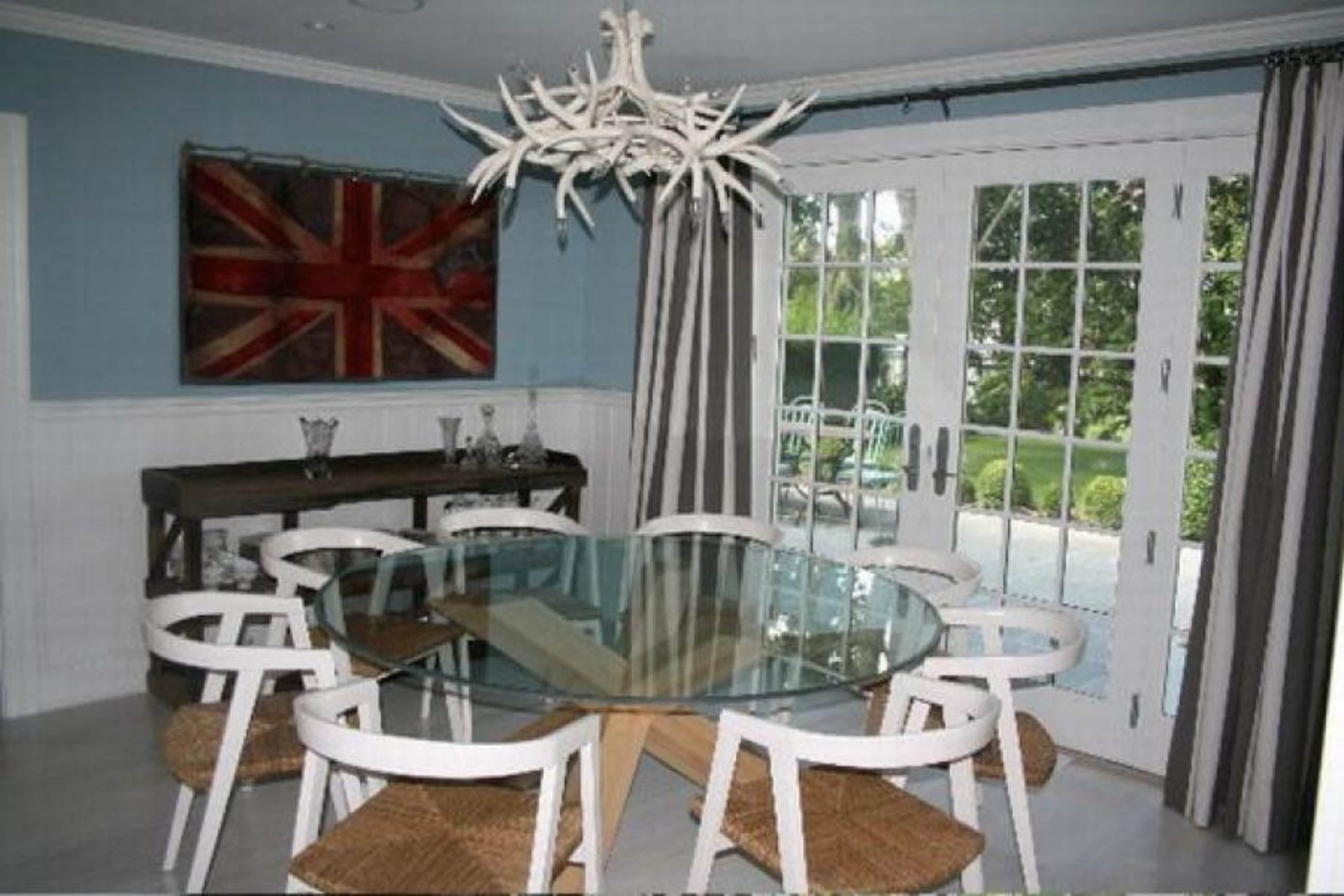 A dining area features a round glass table surrounded by white chairs with woven seats, alongside a British flag artwork and large glass doors leading to a garden.