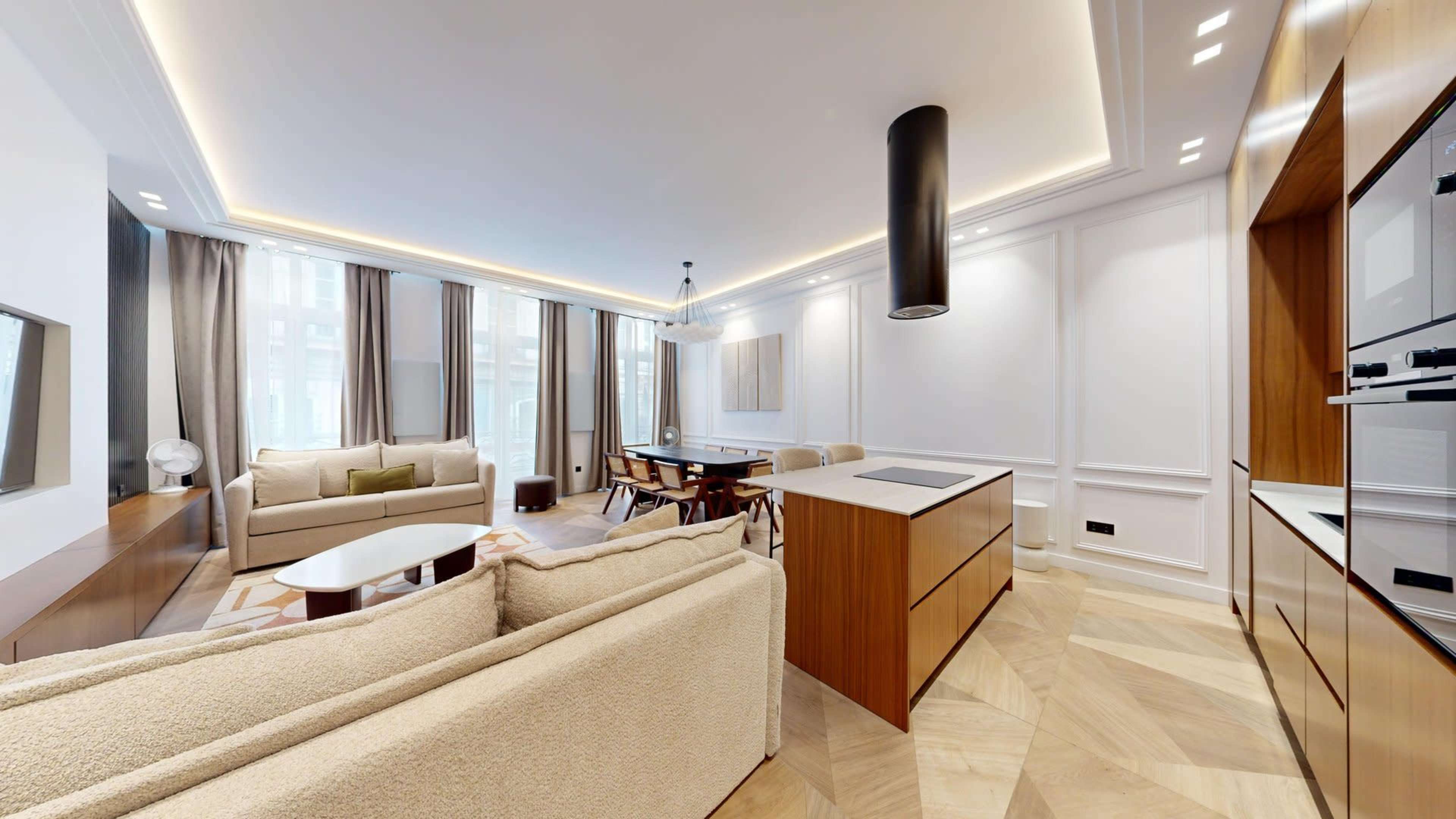 The image shows a modern living room with a large seating area, a dining table, and an open kitchen featuring a central island.