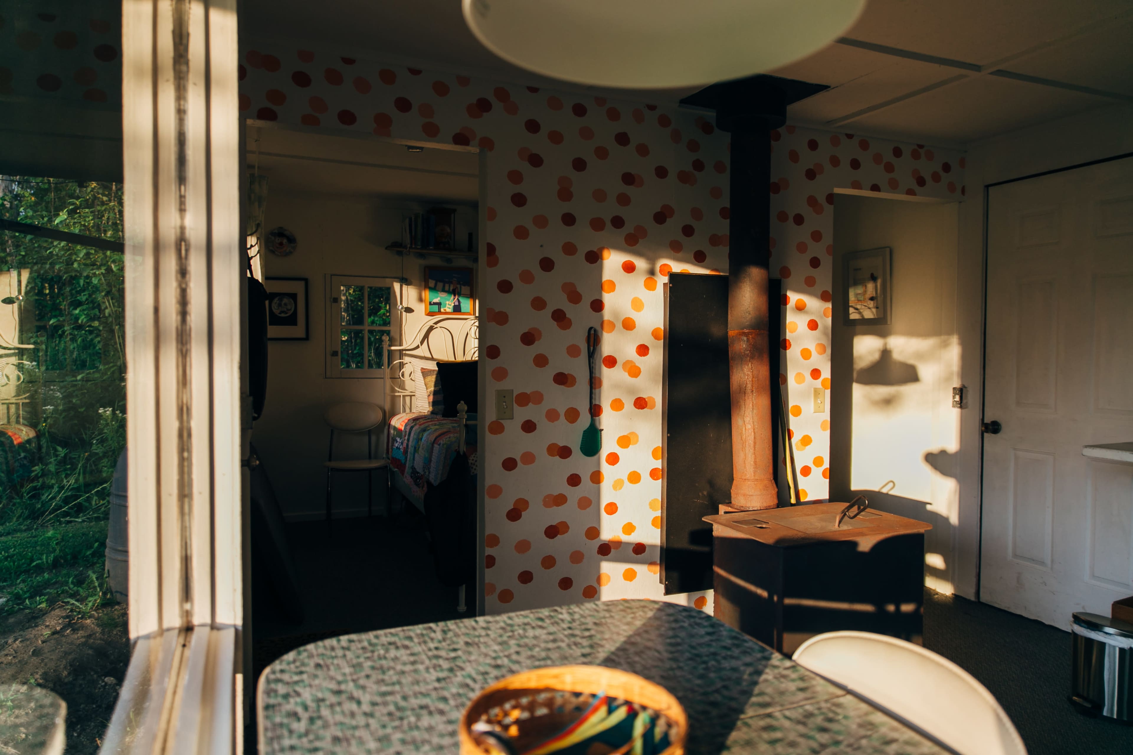 A sunlit kitchen features a table with a bowl, colorful polka-dot wallpaper, and a wood-burning stove in the corner.