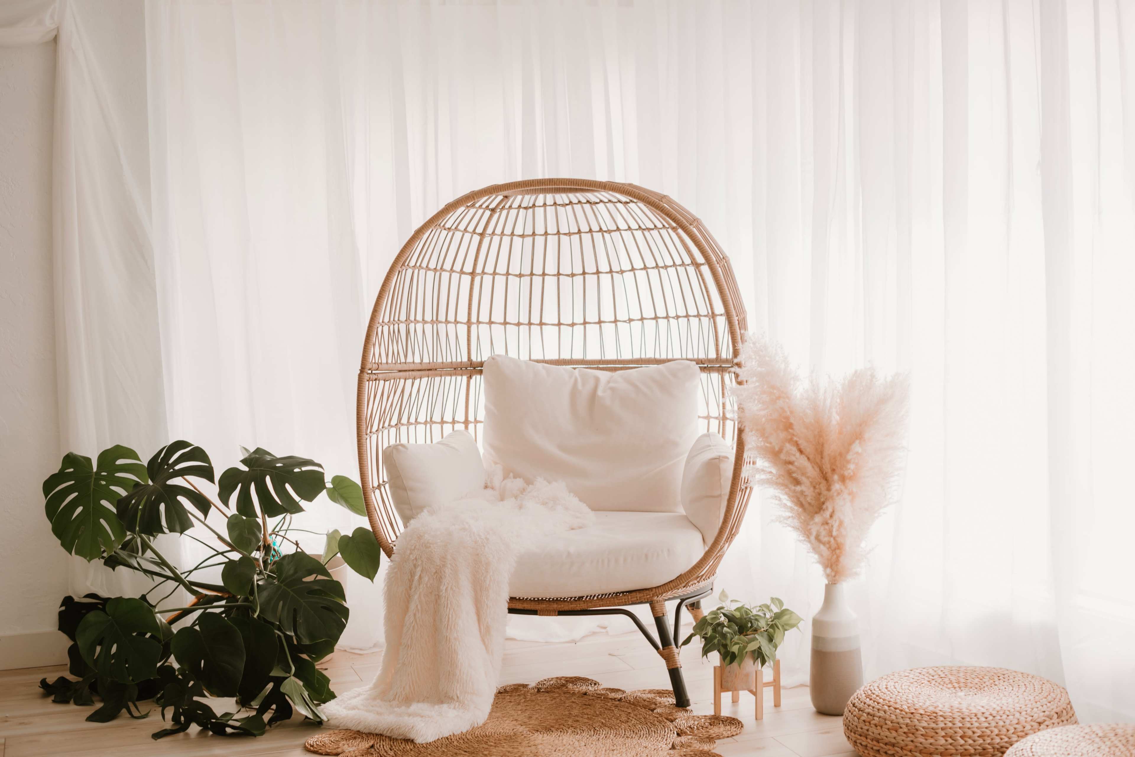 A rattan lounge chair with white cushions is positioned near a large window, surrounded by plants and decorative elements.
