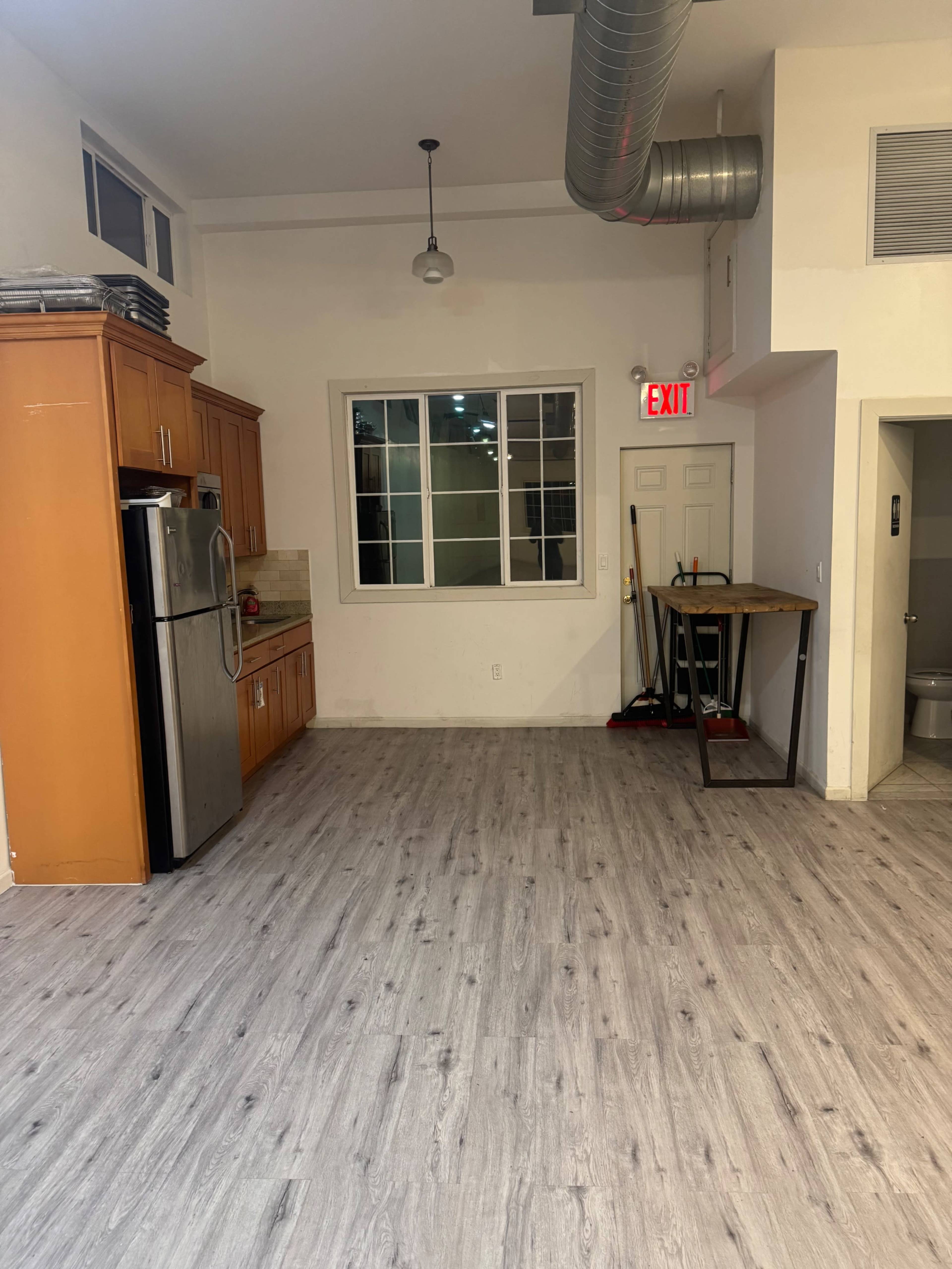 The image shows a kitchen area with wood cabinets and a stainless steel refrigerator, adjacent to a spacious, empty room with light-colored laminate flooring.