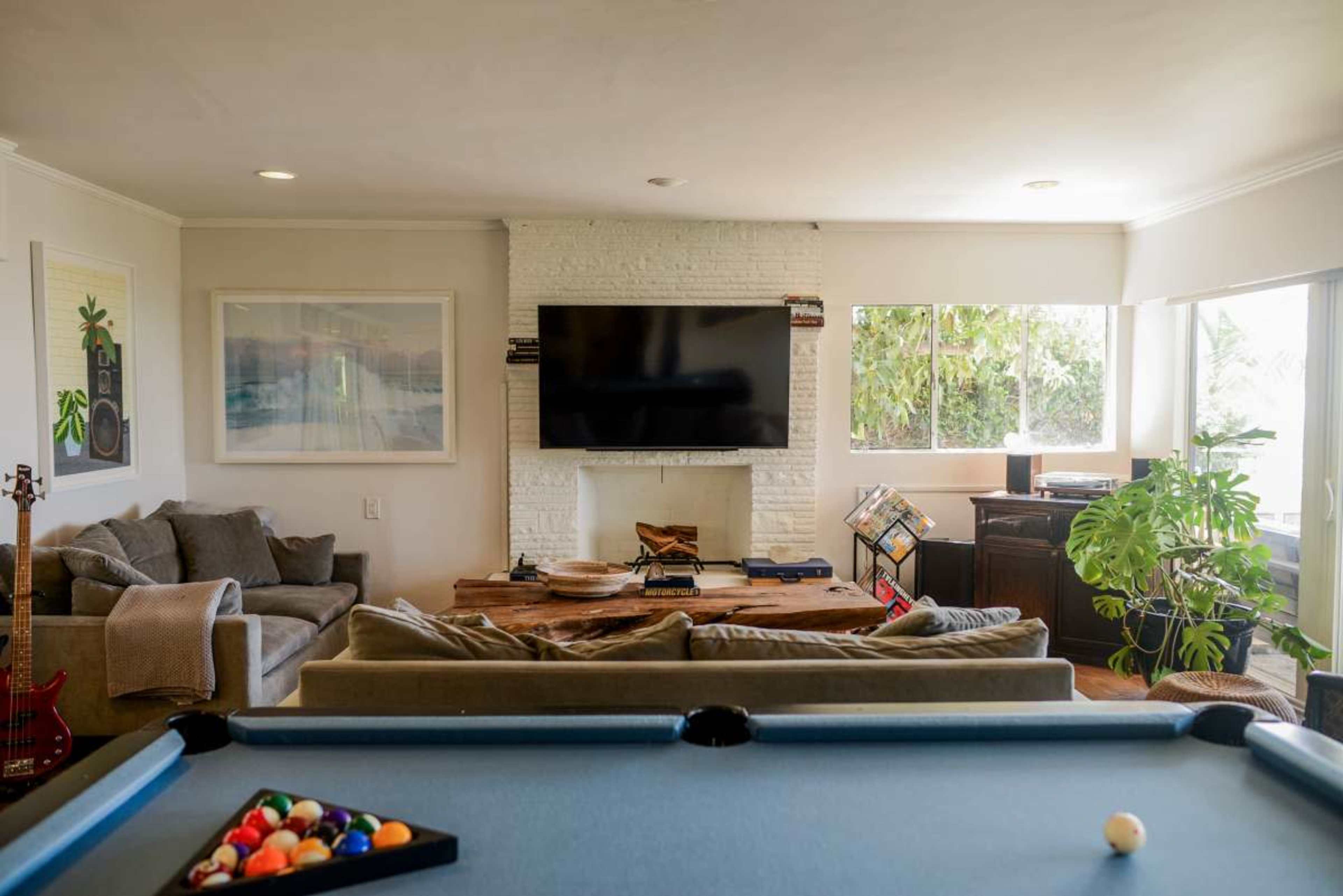 The image shows a living room with a pool table in the foreground, a large TV mounted on a white brick wall, and a cozy seating area with a couch and coffee table surrounded by windows and plants.