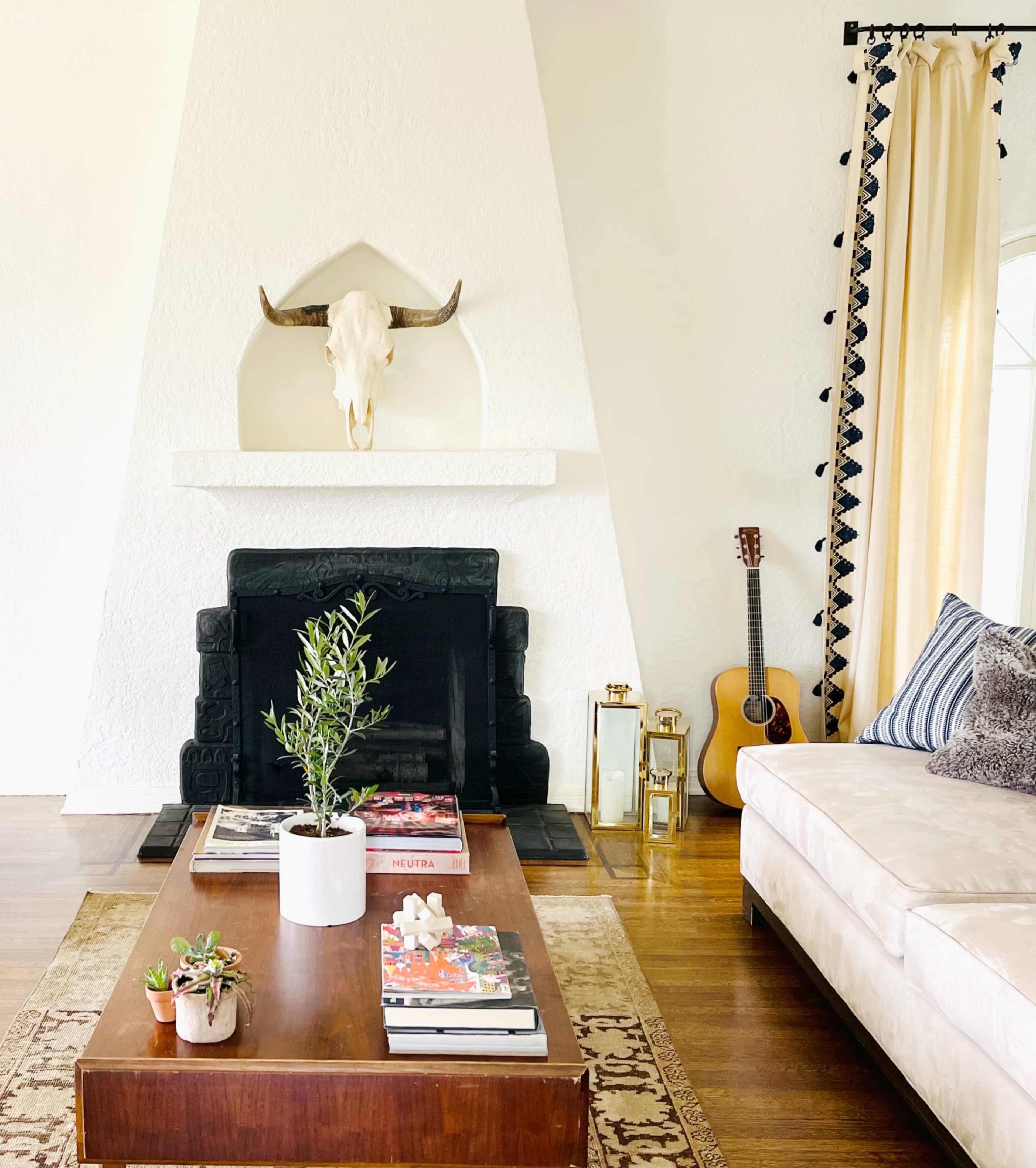 A living room featuring a fireplace with a decorative bull skull above it, a wooden coffee table with magazines, a potted plant, and a guitar leaning against the wall.