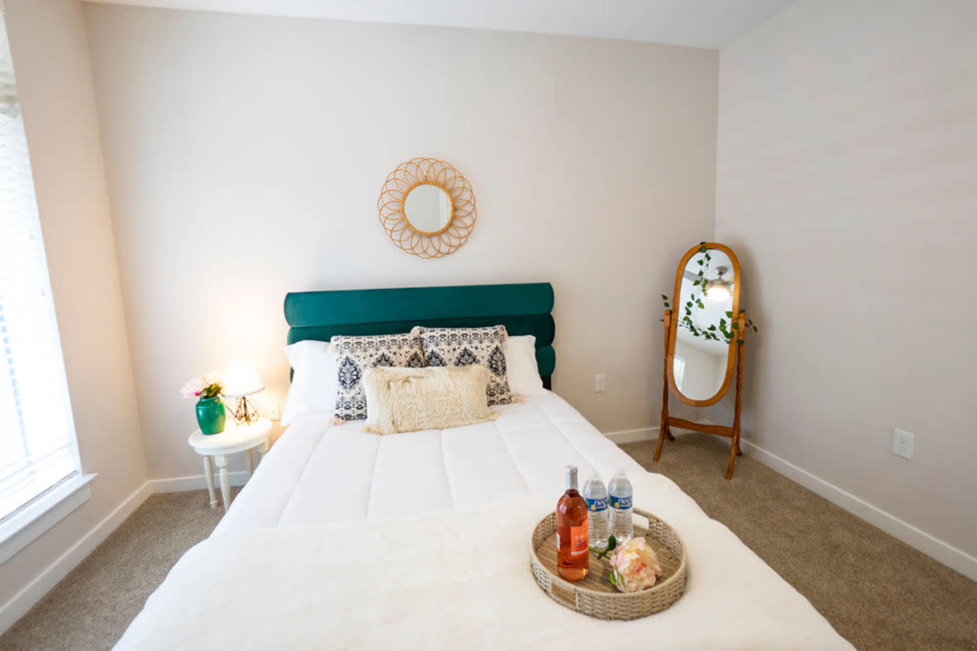 A neatly arranged bedroom featuring a green-headboard bed, a round wall mirror, and a tray with two bottles and a floral arrangement on the bed.