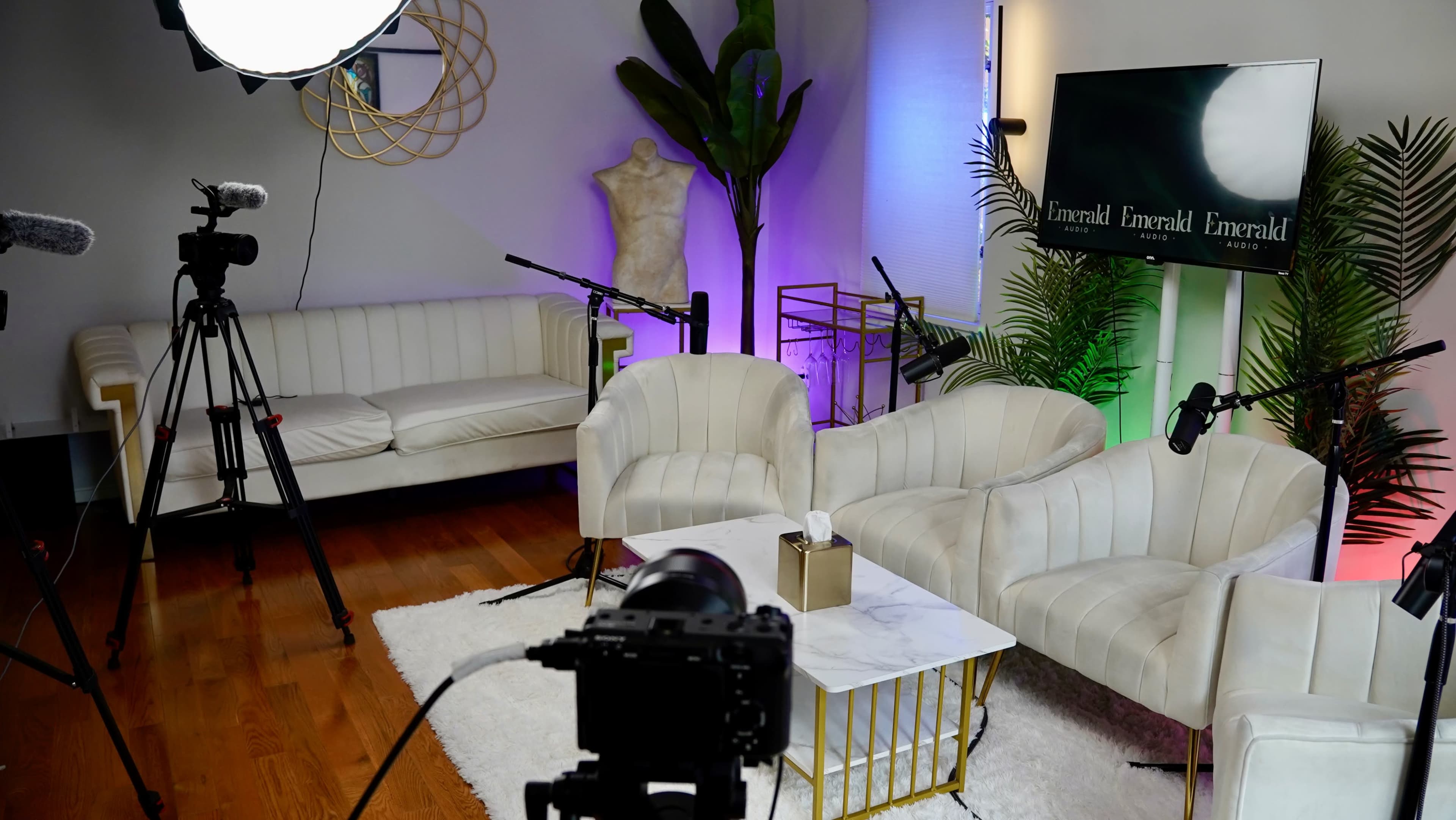 A modern studio setup with white chairs, a marble coffee table, a camera on a tripod, and colorful lighting accents.