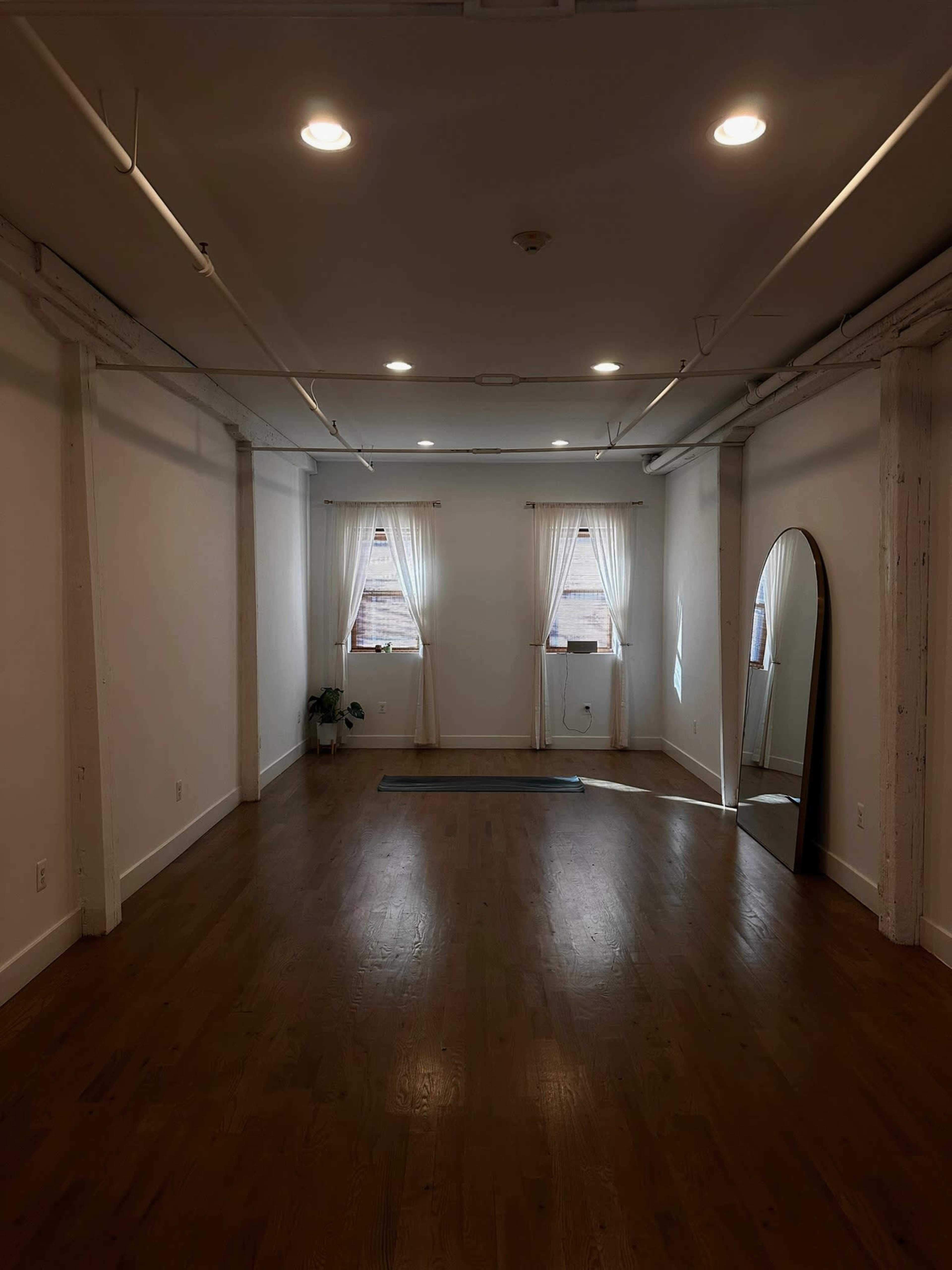 The image shows a spacious, well-lit room with wooden floors, two windows draped with sheer curtains, and a large mirror mounted against a wall.