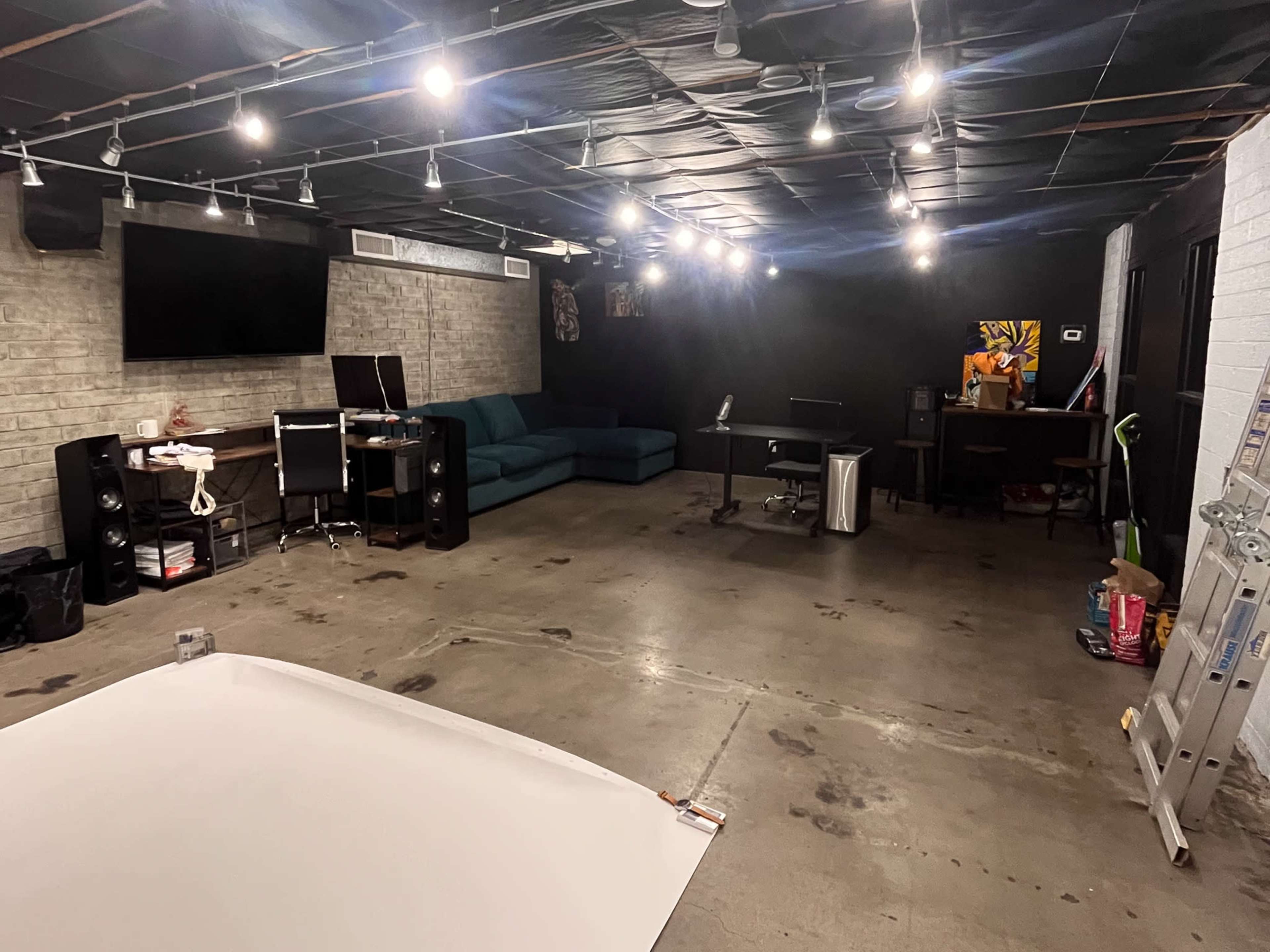 A spacious room with a concrete floor, featuring a green sofa, a desk with a computer, and various audio equipment, all illuminated by overhead track lighting.