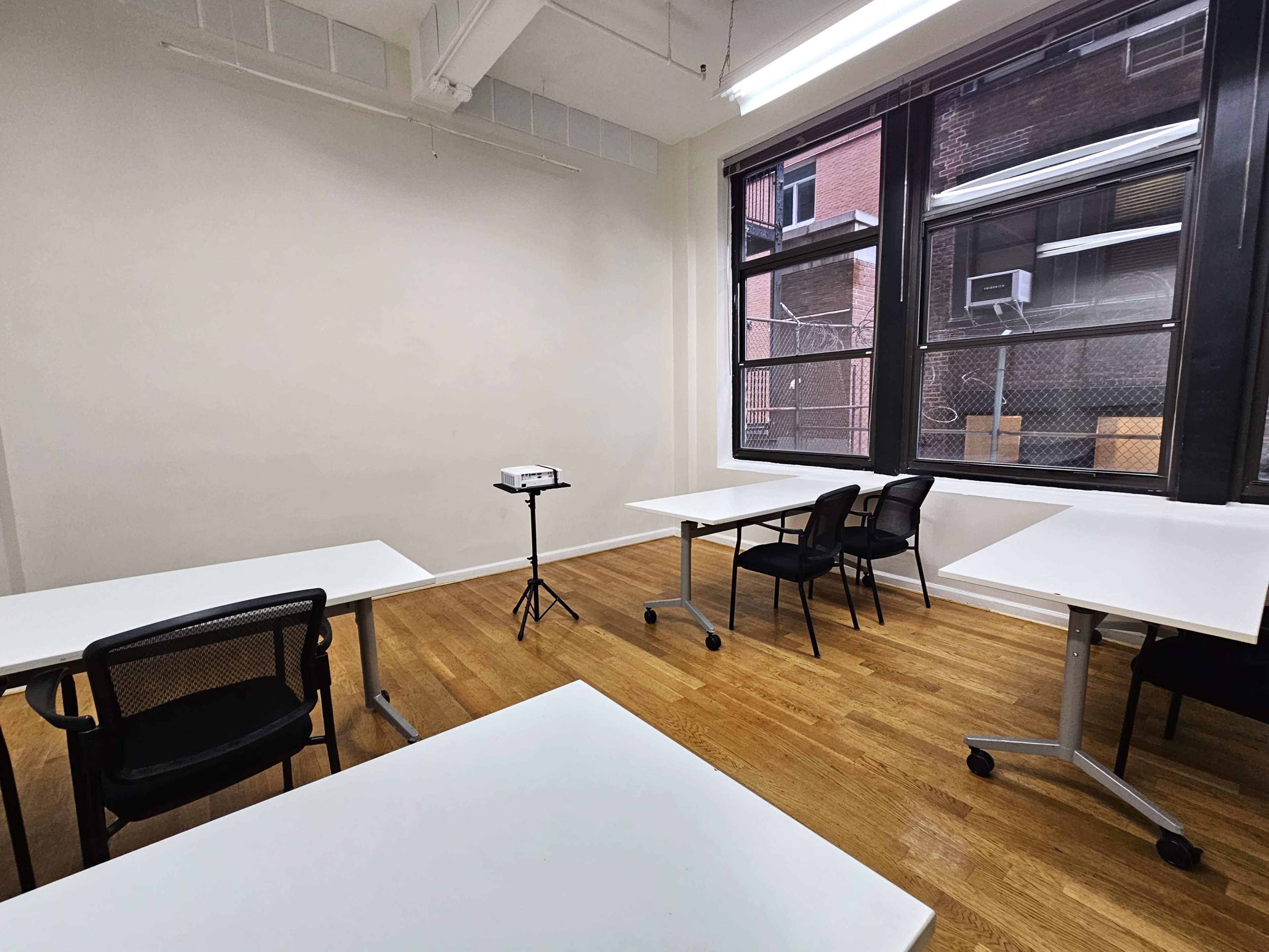 A spacious, empty classroom features white tables and black chairs, with a projector on a stand in the center.