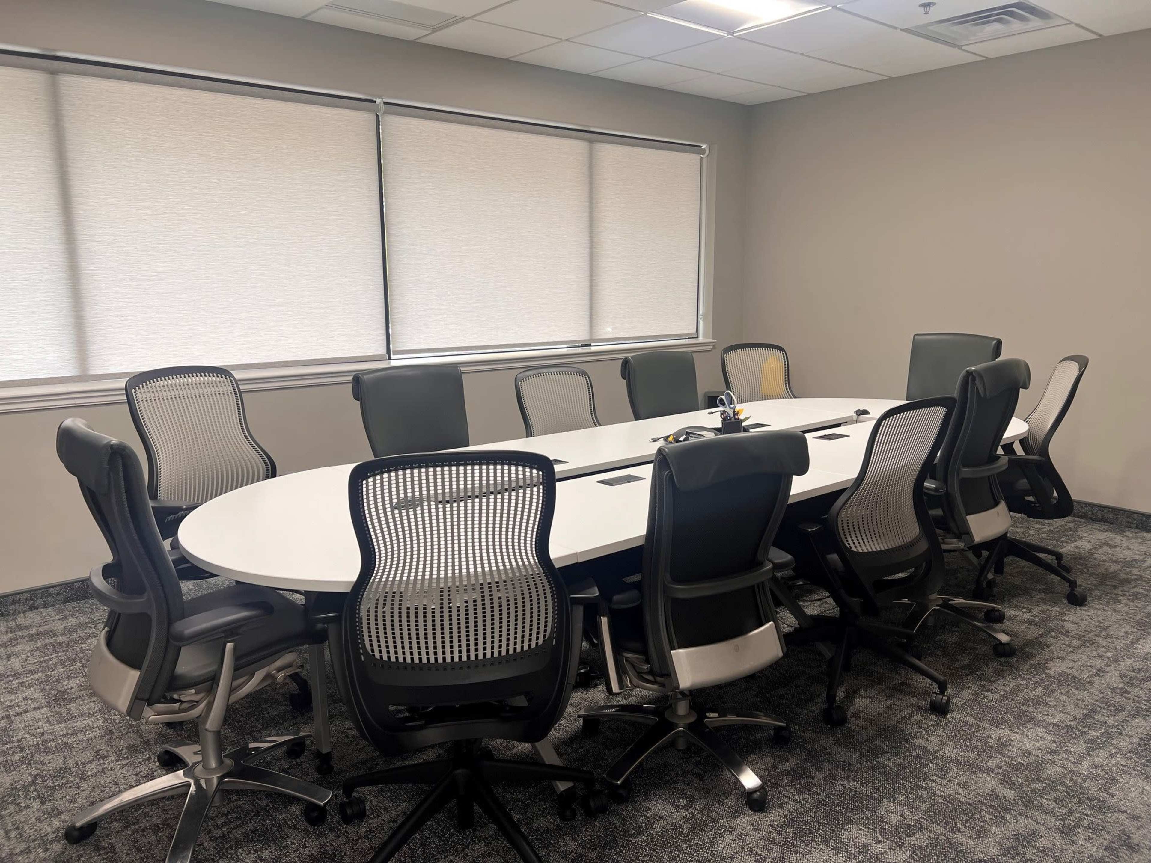 A large, oval conference table is surrounded by ten office chairs in a well-lit meeting room with window shades drawn.