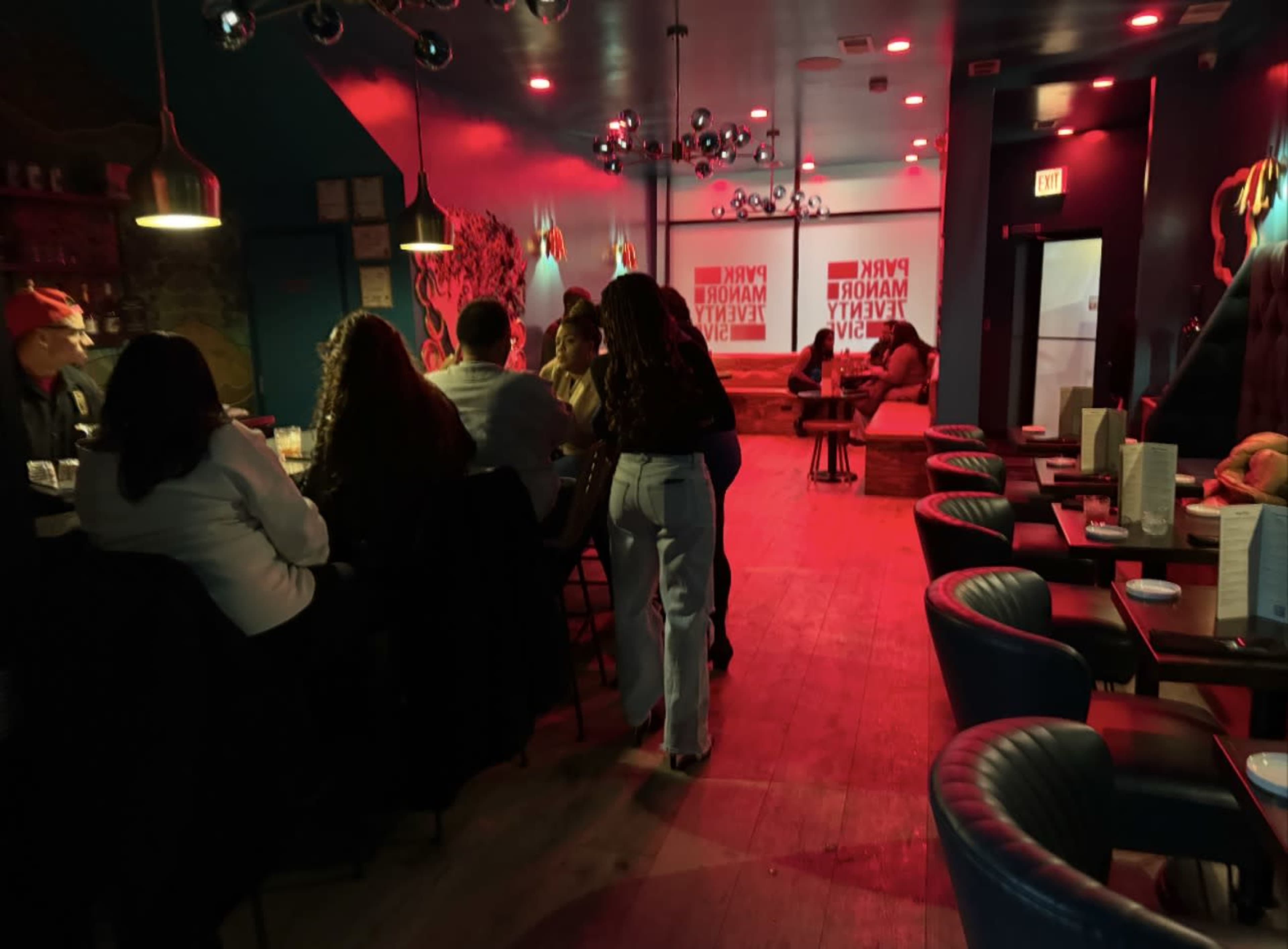 A dimly lit lounge features patrons seated at tables and a bar, with colorful lighting and decorative elements creating a vibrant atmosphere.