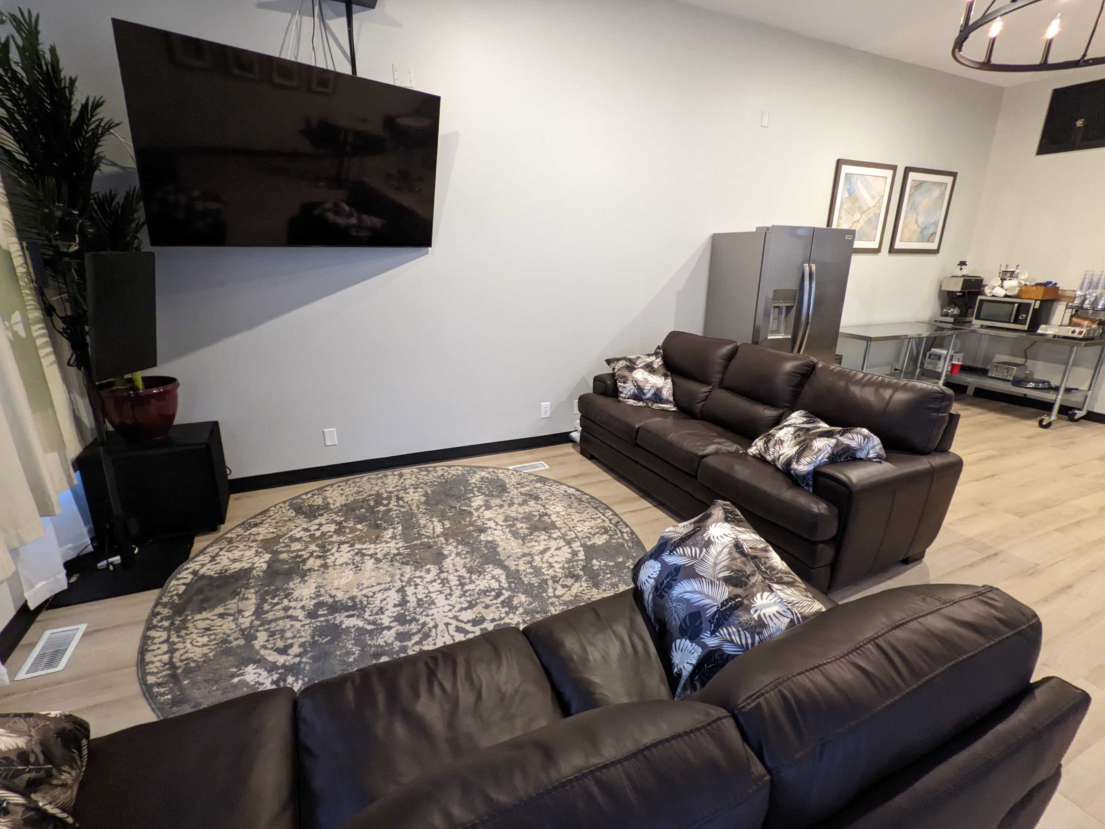 A modern living room with two brown sofas, a round area rug, a wall-mounted television, and a stainless steel refrigerator in the background.