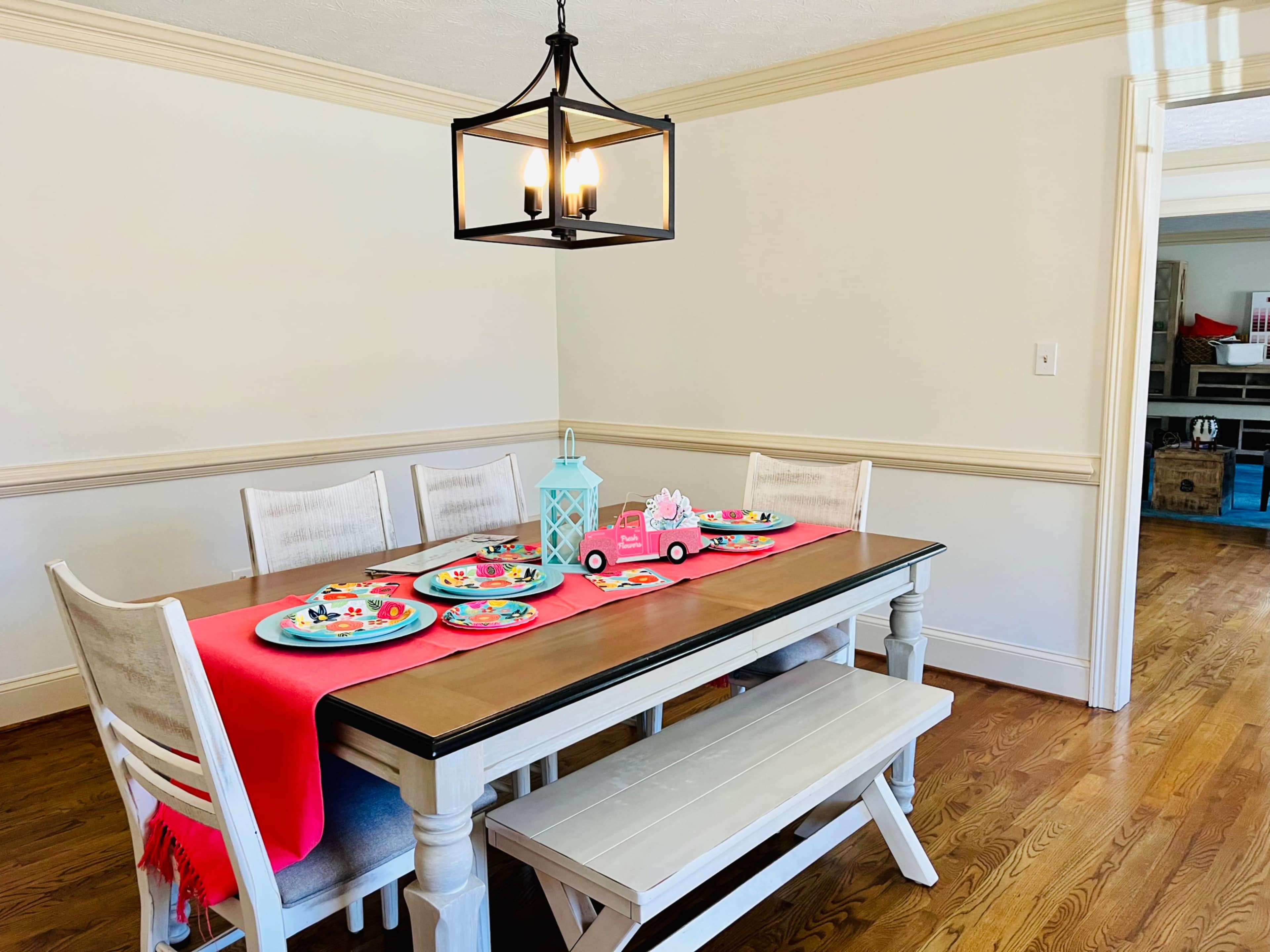 A wooden dining table is set with colorful plates and a pink decorative truck under a pendant light in a bright room.