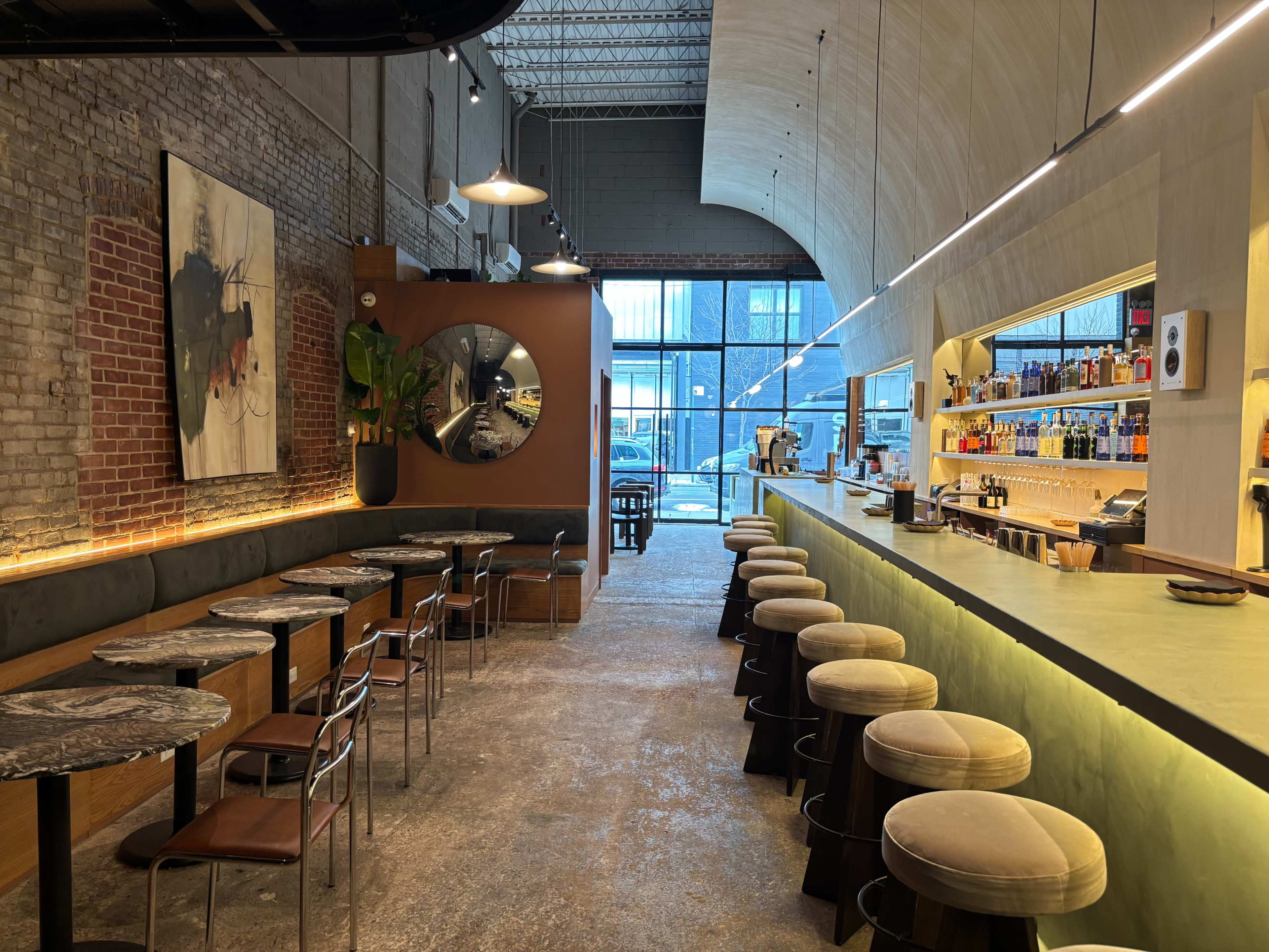 The image shows a modern café interior with a long bar, seating along the wall, large windows letting in natural light, and industrial elements like exposed brick and high ceilings.