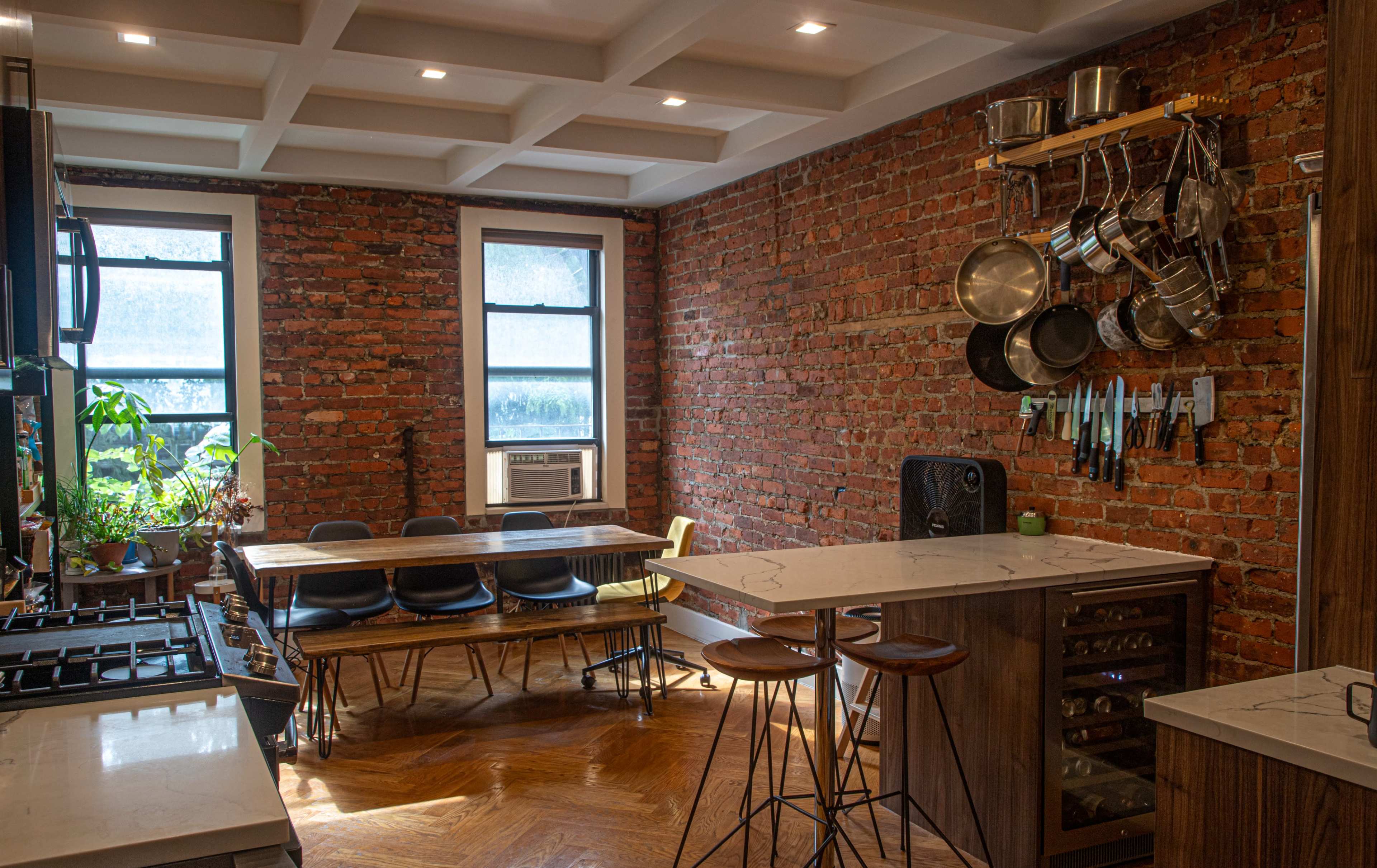 A modern kitchen features exposed brick walls, a large wooden table, and stainless steel cookware hanging above the countertop.