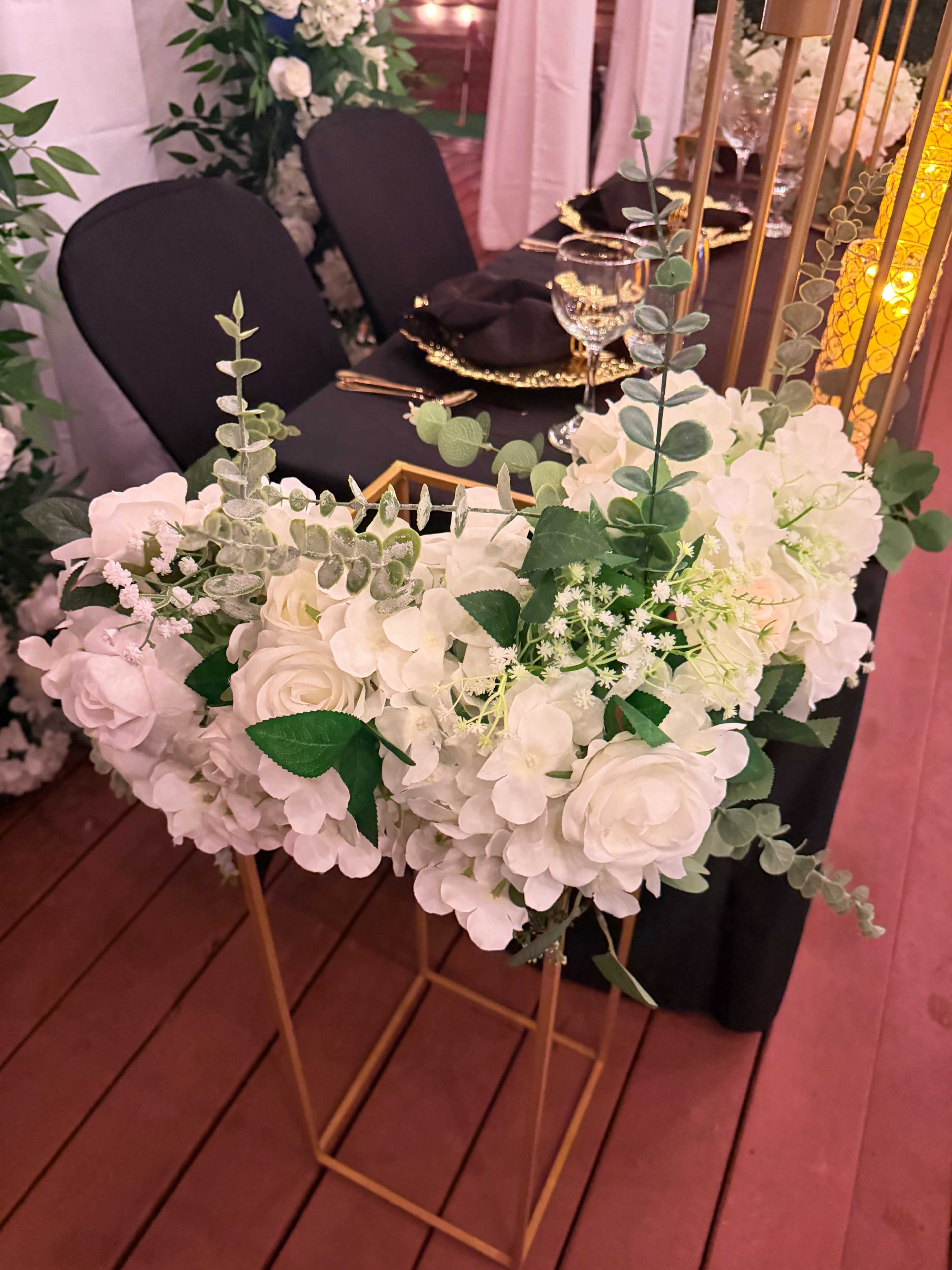 The image shows a decorative floral arrangement featuring white flowers and green foliage, positioned on a gold metal stand at an event setting.