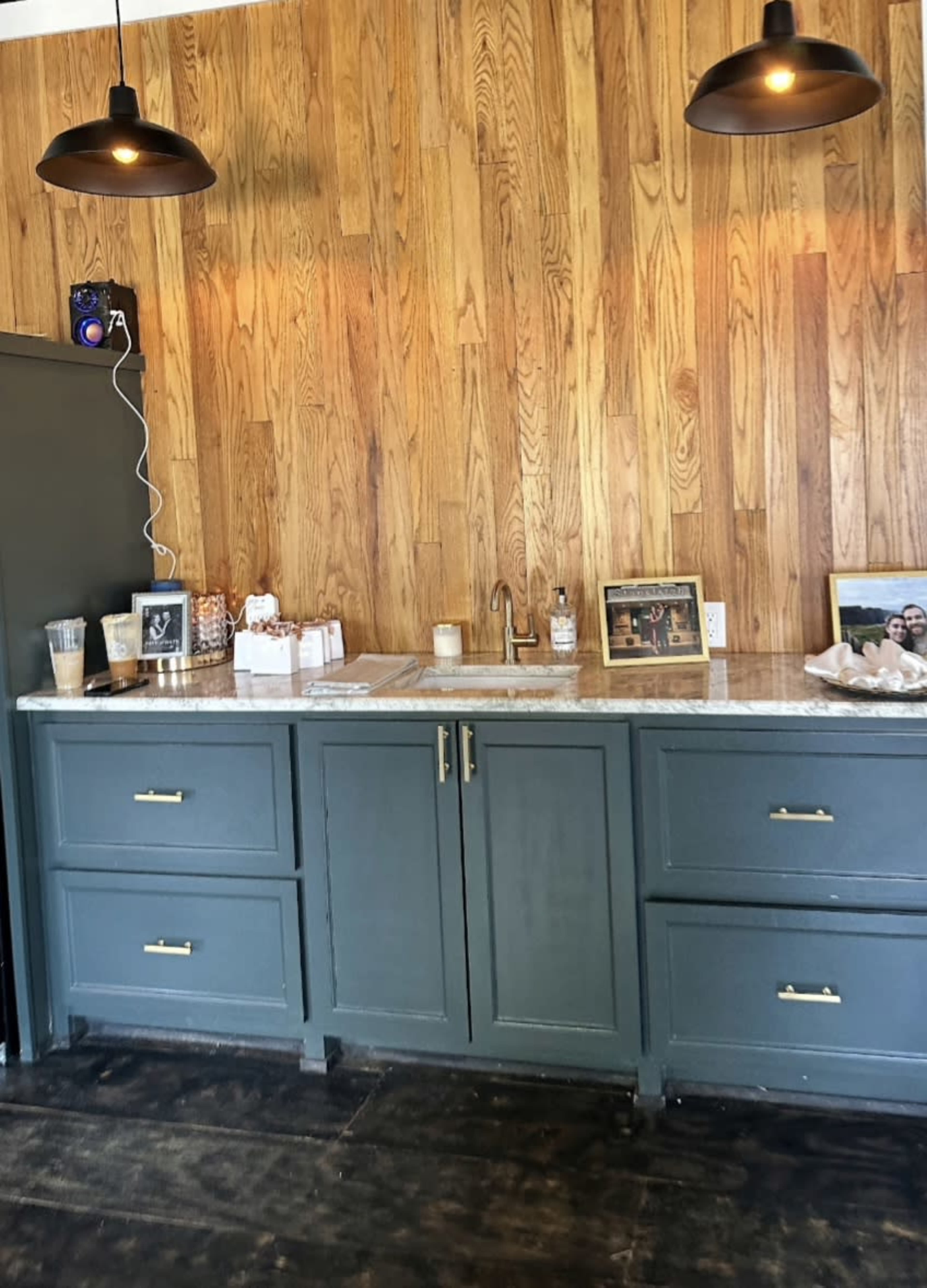 The image shows a wooden-paneled wall with a dark green cabinet containing a sink, counter space, framed photos, and decorative items under two pendant lights.