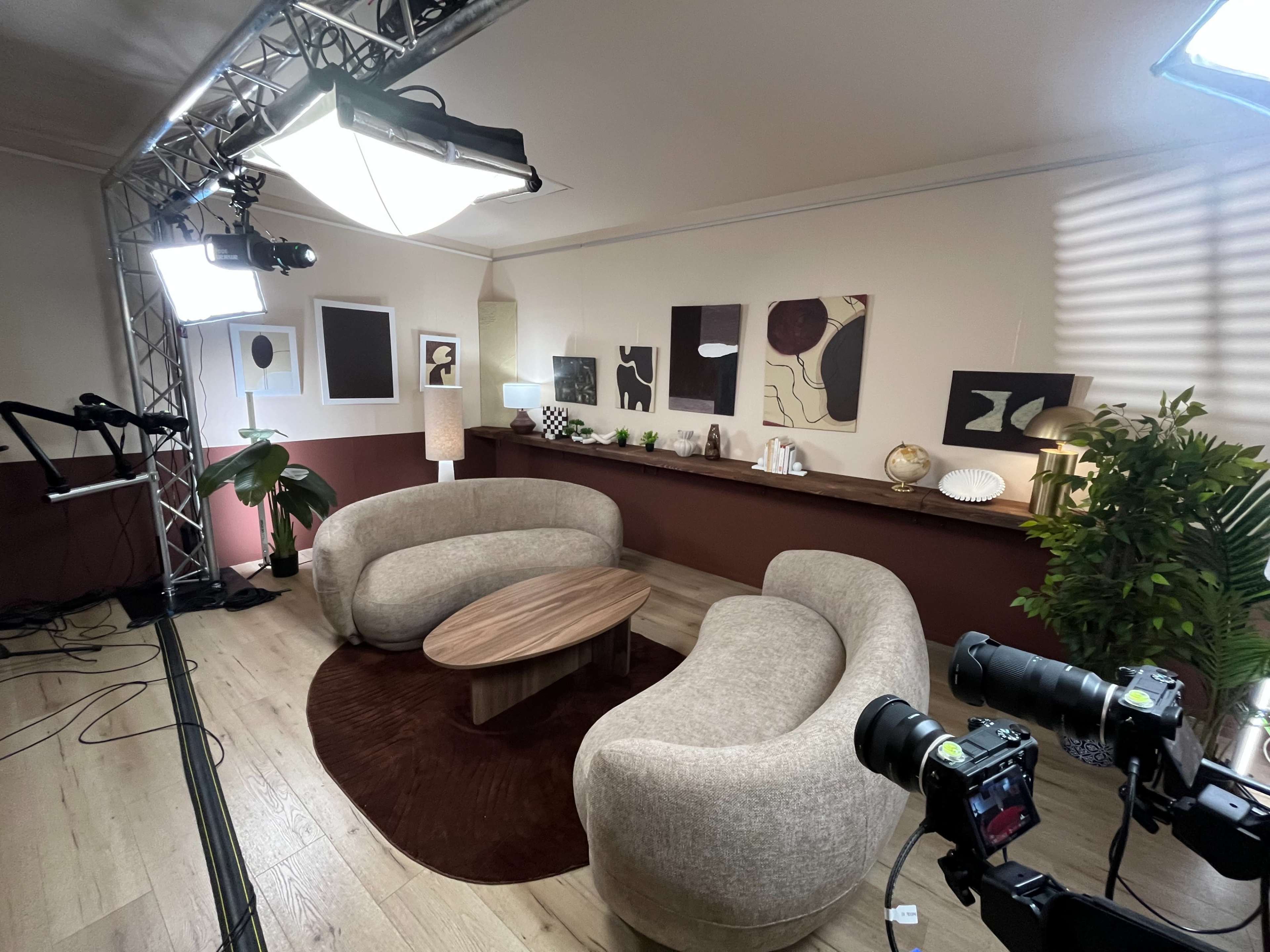 The image shows a modern, well-lit studio set with curved sofas, a round coffee table, and artistic wall decorations.