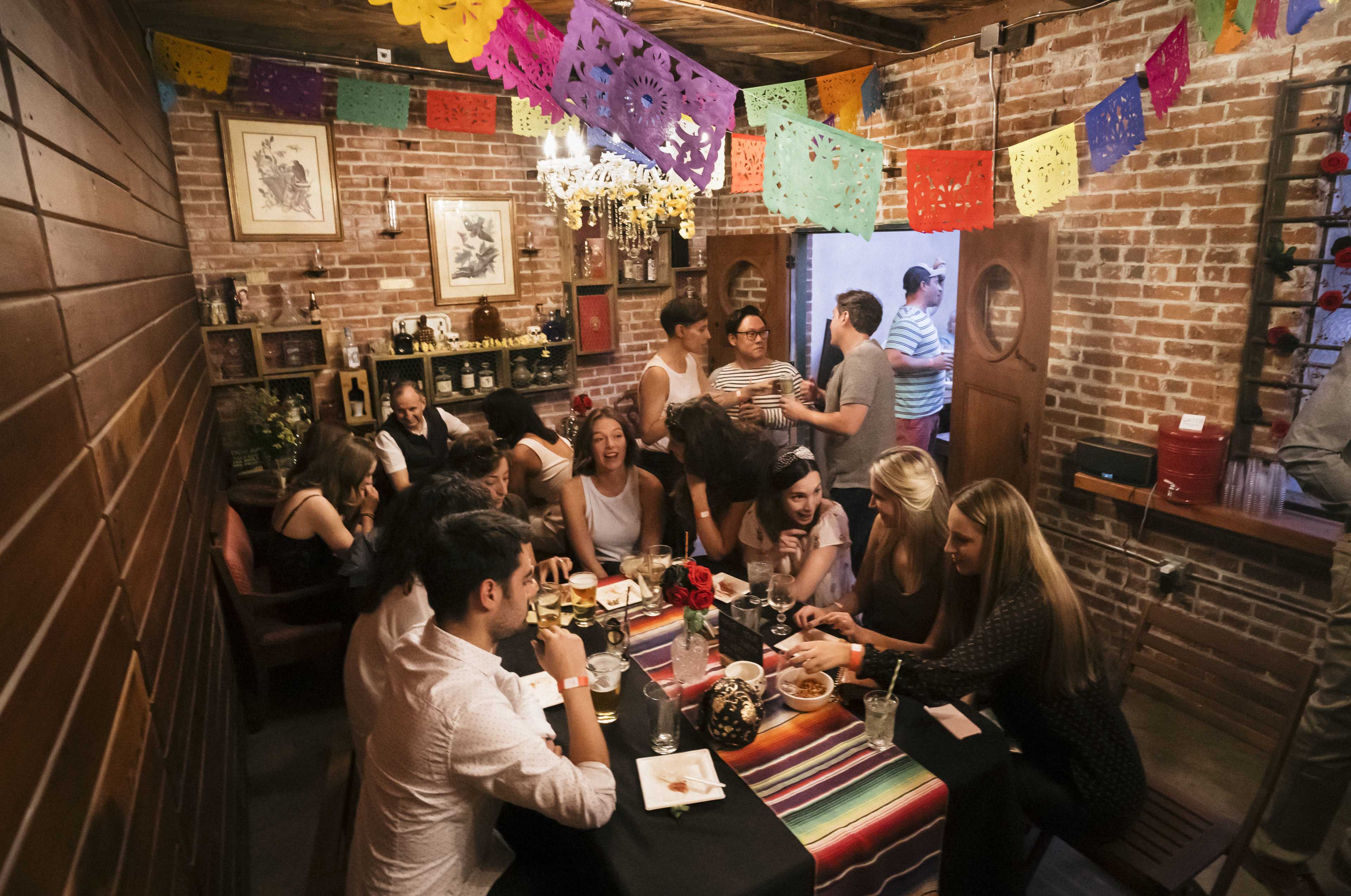 A group of people gathers around a table in a cozy restaurant adorned with colorful papel picado decorations and exposed brick walls.