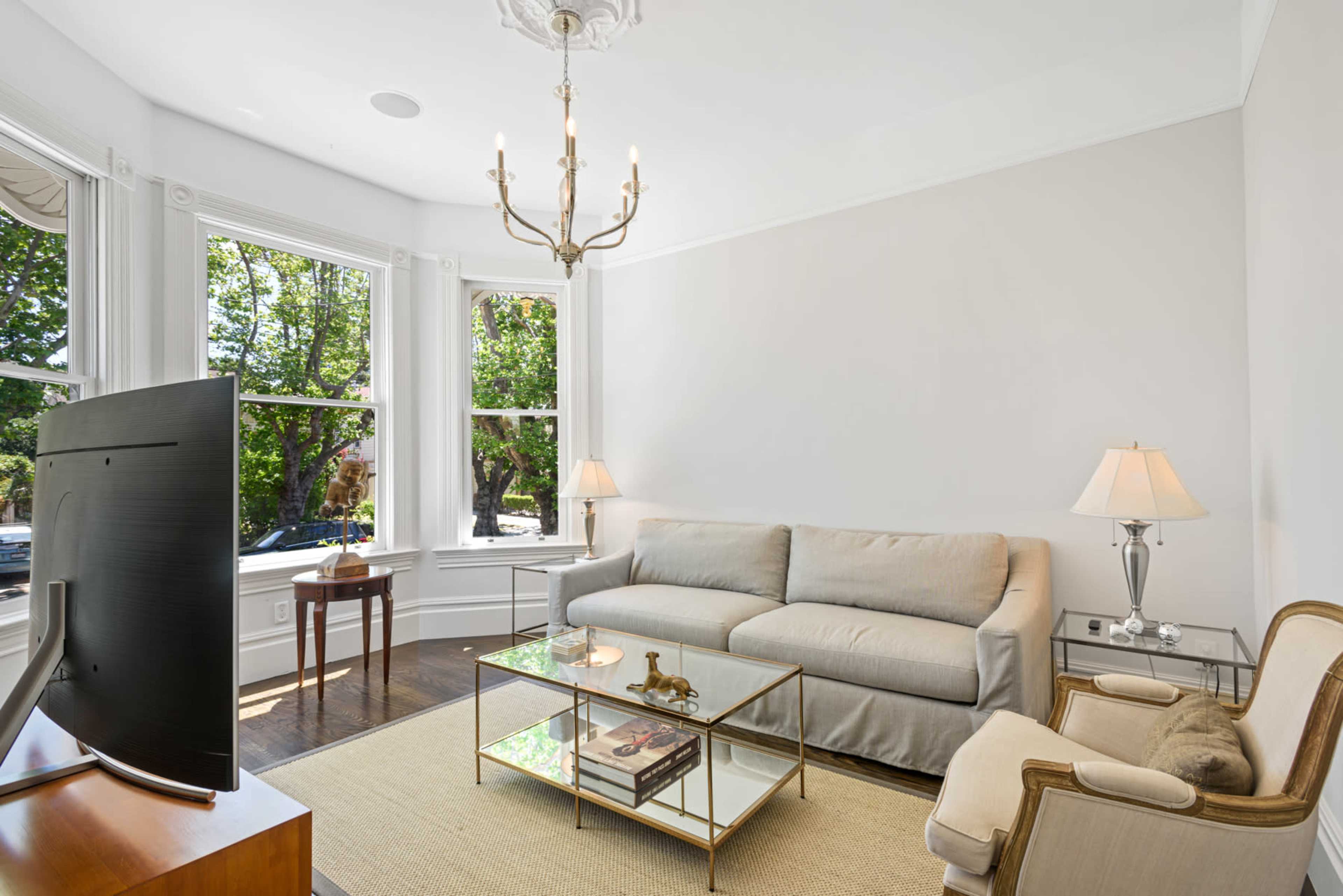 A bright living room features a beige couch, a glass coffee table, and two lamps beside a window with views of trees outside.