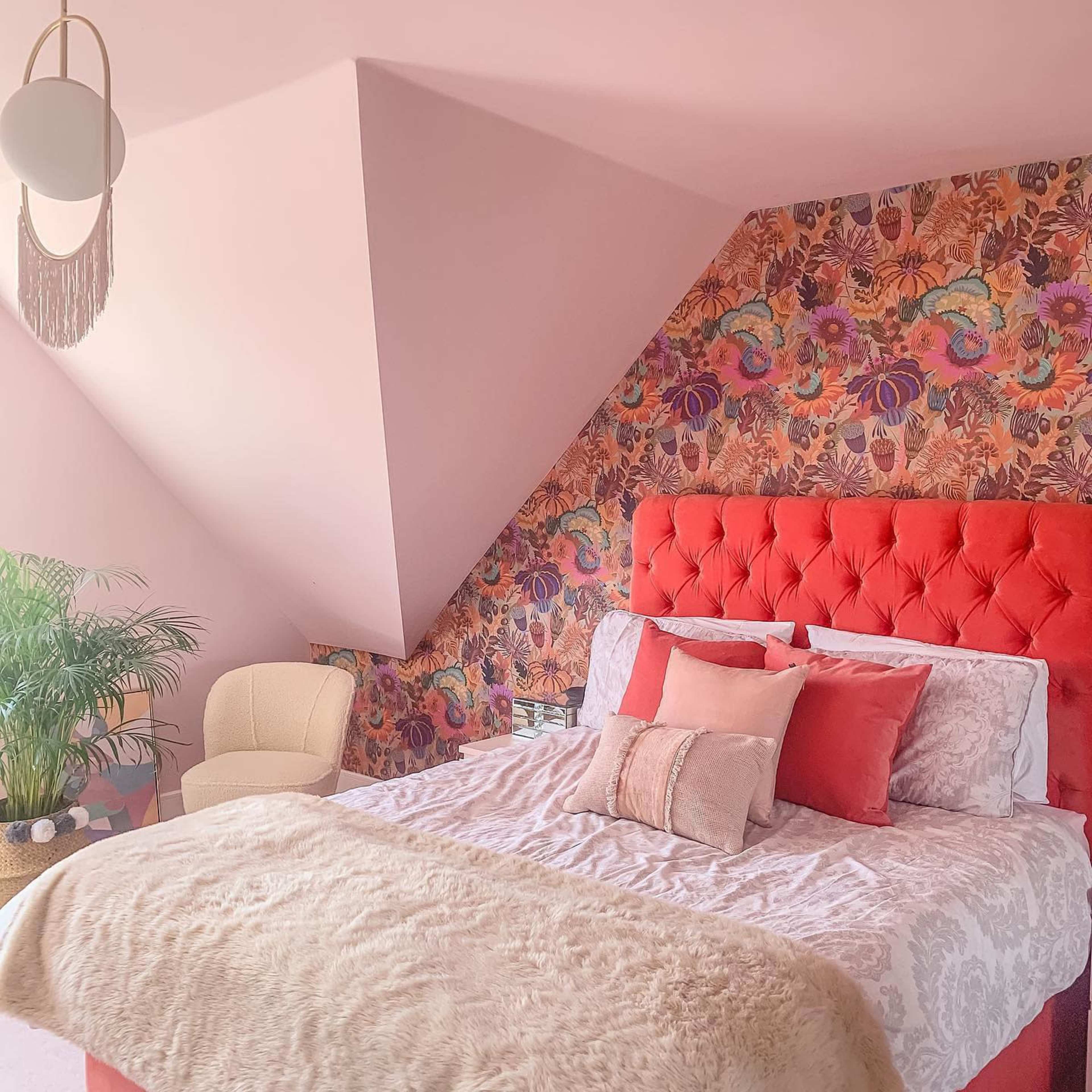 A bedroom with a pink and floral patterned accent wall, a red upholstered headboard, a white bedspread, and a cozy armchair.