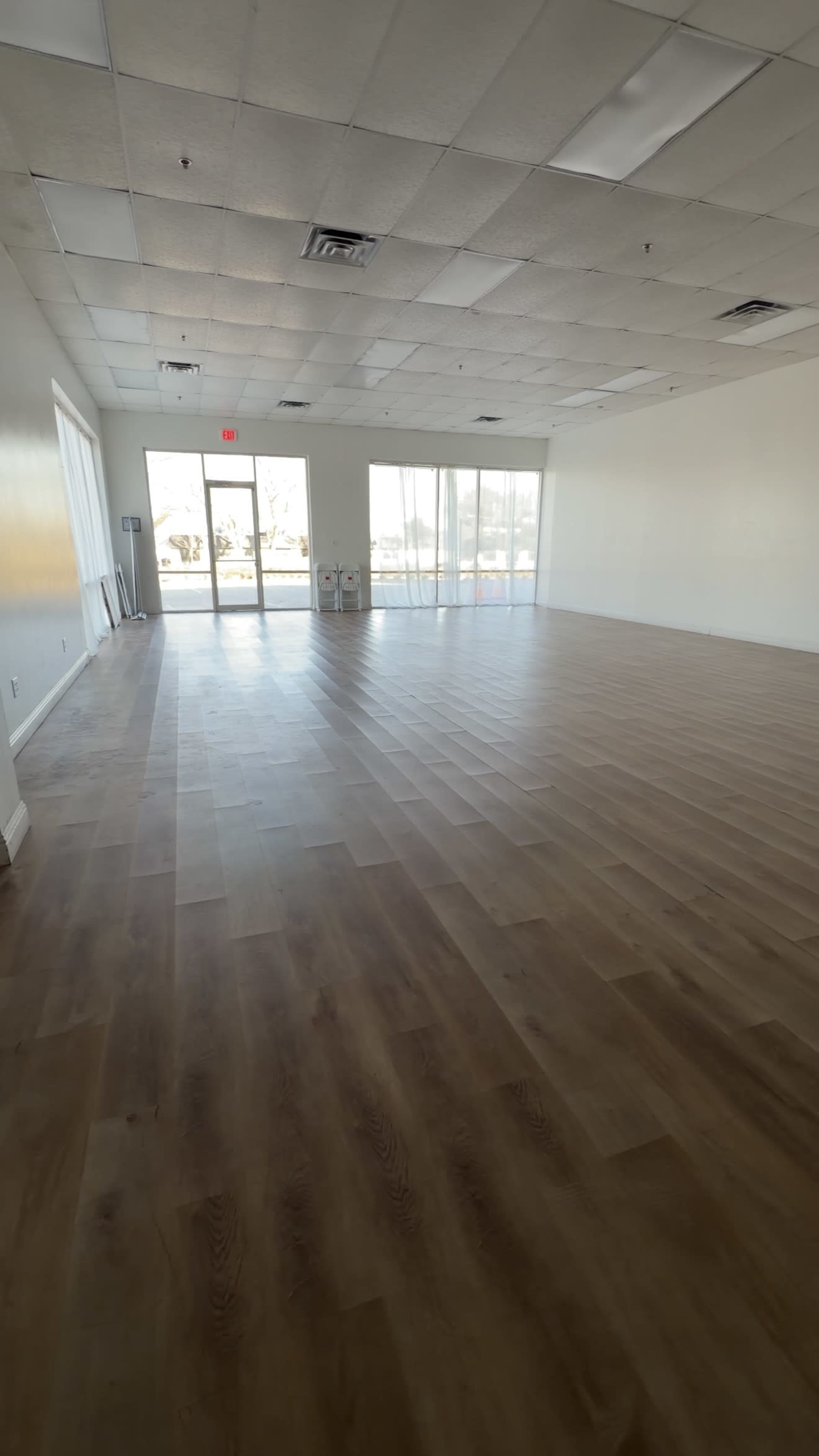 The image shows a spacious, empty interior room with wooden flooring and large windows that let in natural light.