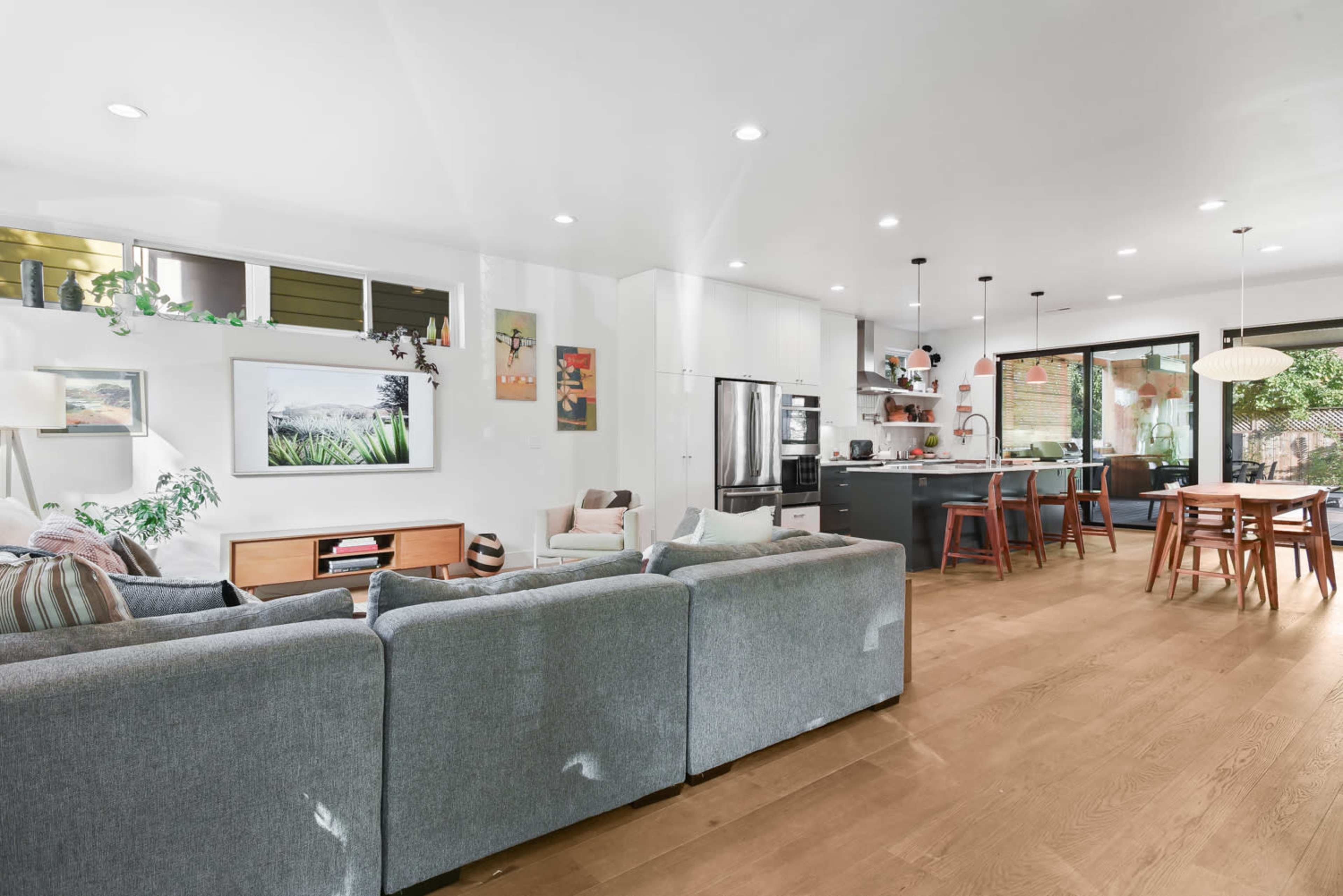 A modern living space features a gray sectional sofa, a dining area with a wooden table, and an open kitchen with black cabinetry and stainless steel appliances.