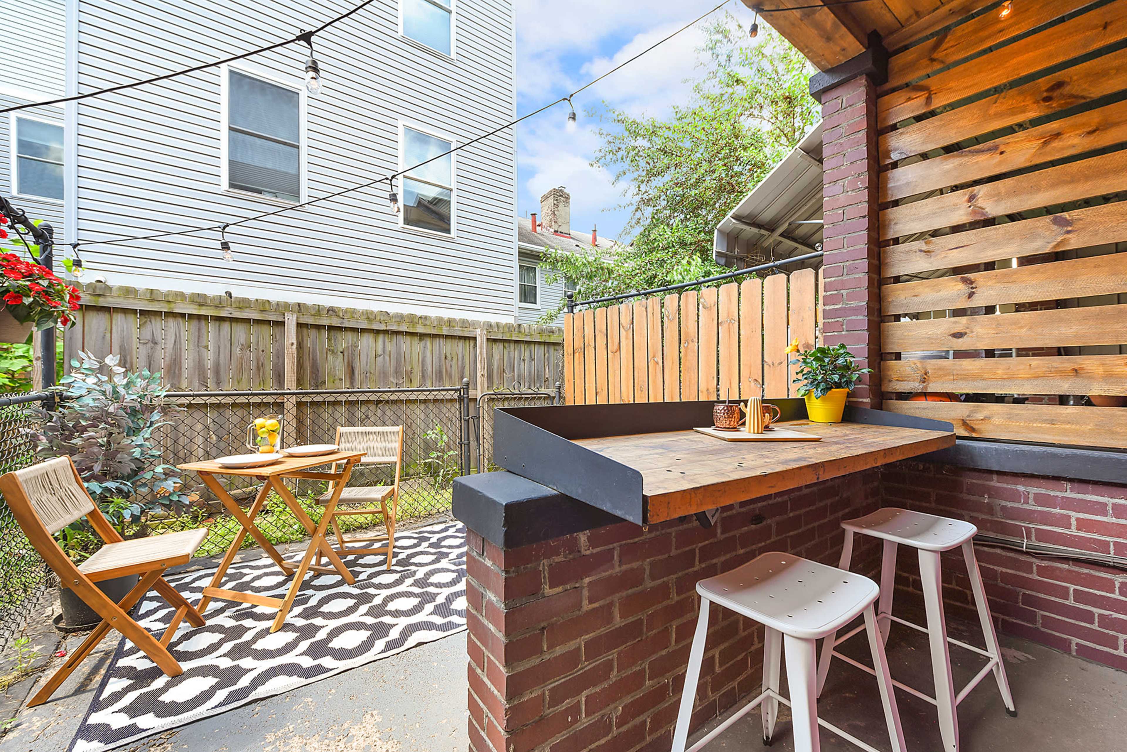 The image shows a small outdoor patio area featuring a wooden bar with stools, a table and chairs, and string lights overhead, surrounded by a fenced yard.