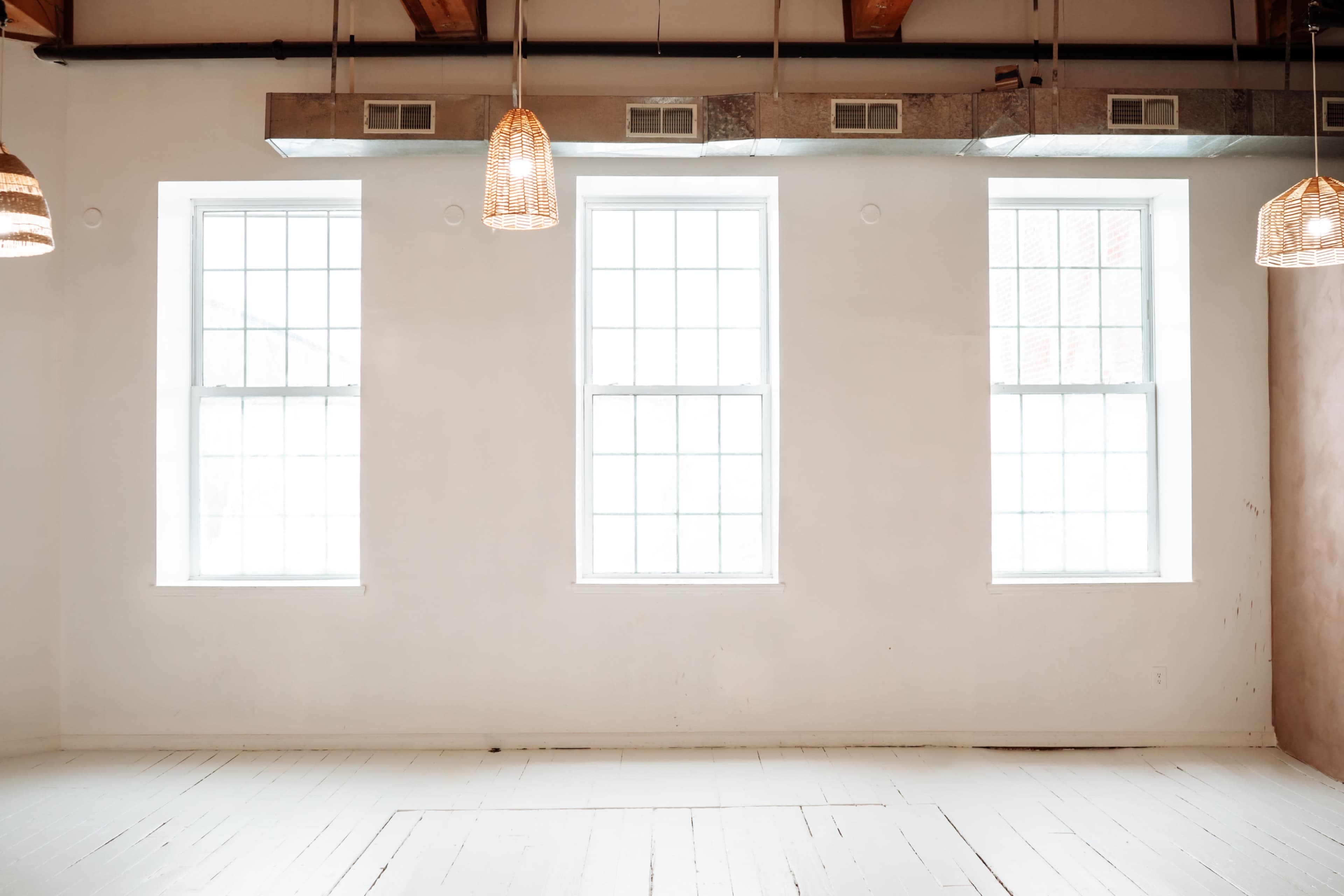 The image shows a minimalist interior space with three large windows and wooden ceiling beams, illuminated by pendant lights.