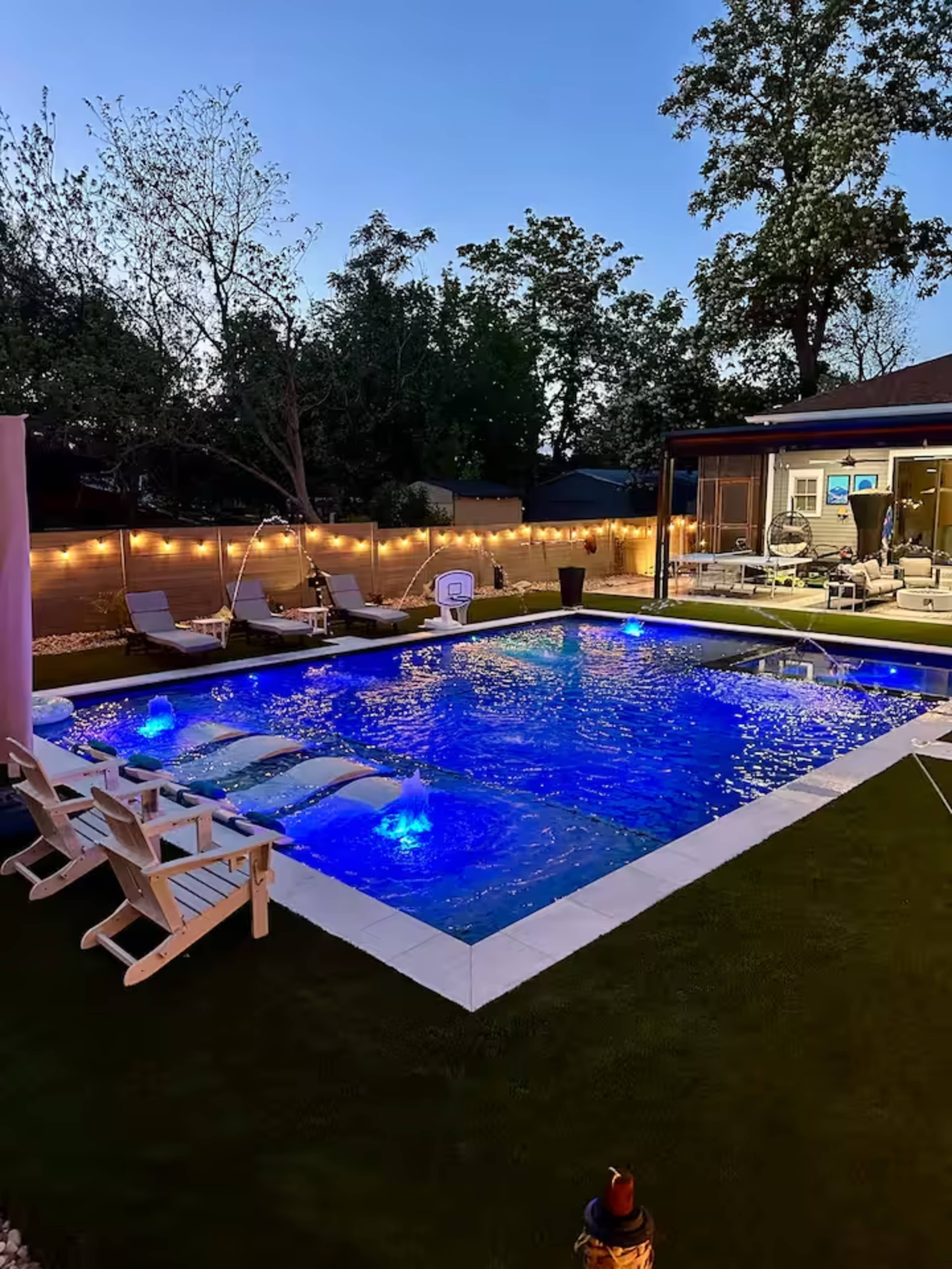 The image shows a well-lit swimming pool surrounded by lounge chairs and greenery, with a modern house featuring large windows in the background.