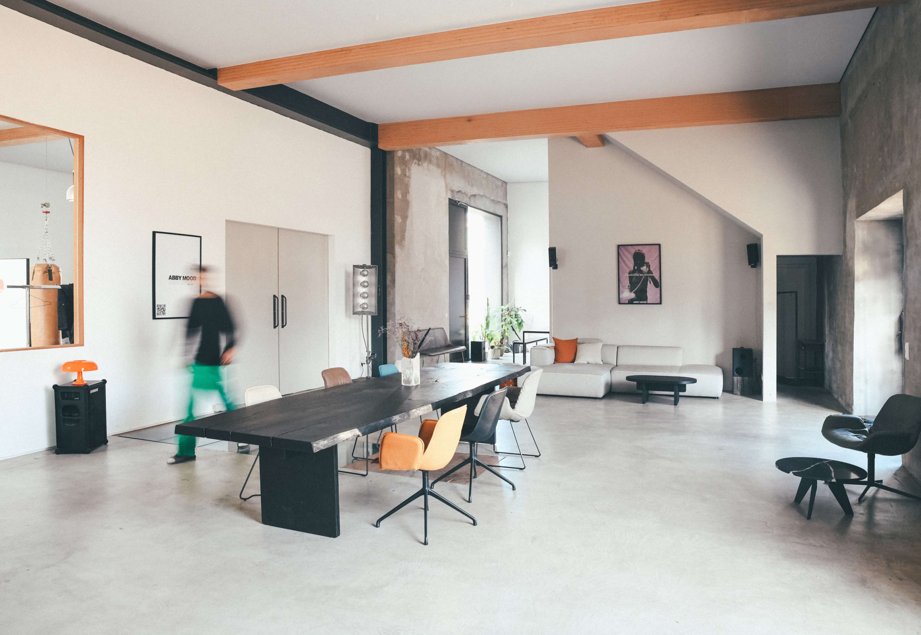 A spacious, modern living area features a long dining table surrounded by various chairs, a sofa in the corner, and large windows that allow natural light to fill the room.