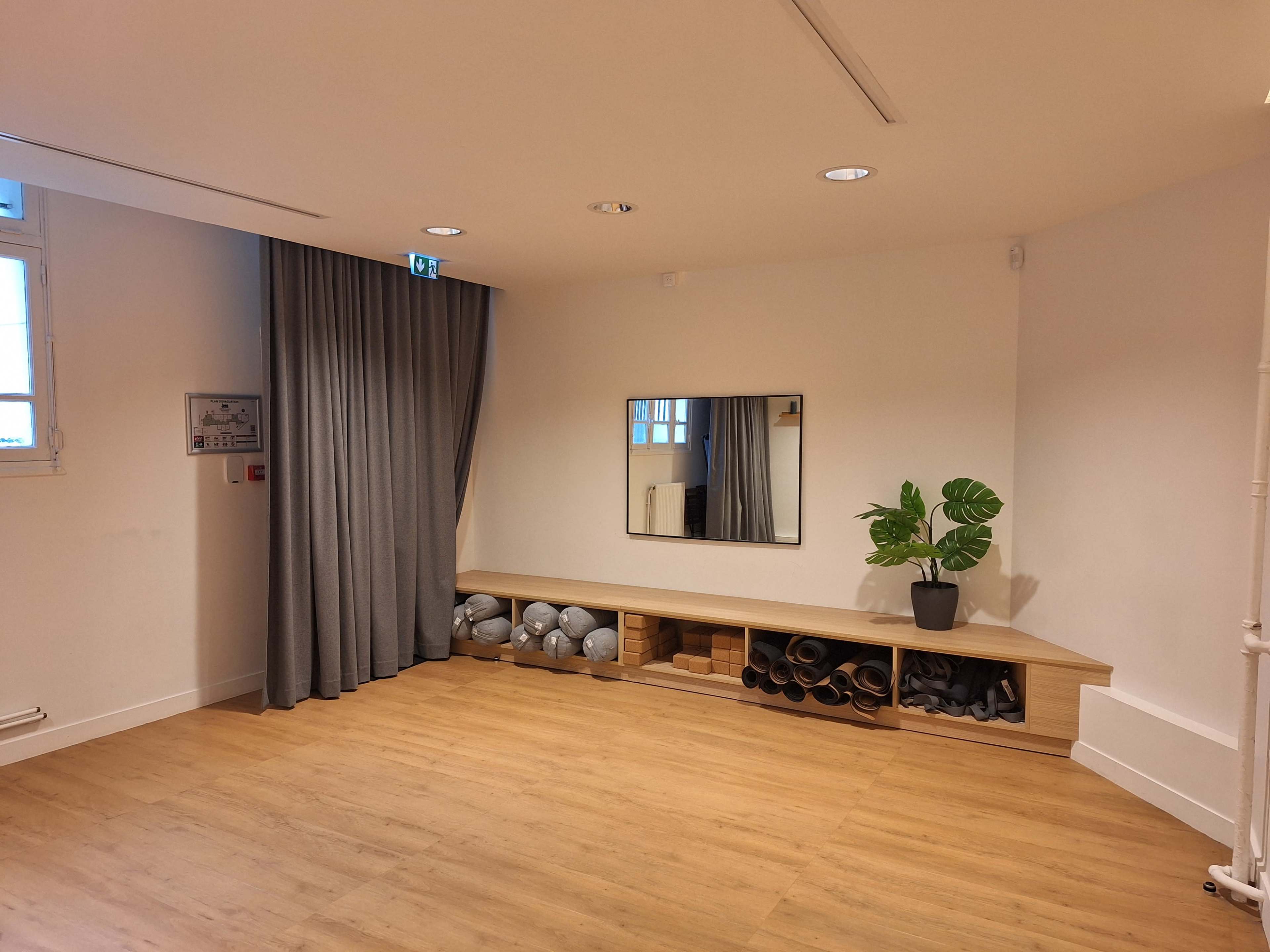 The image shows a spacious, minimalistic room with wooden flooring, a large mirror on the wall, and a bench with rolled mats and storage underneath.