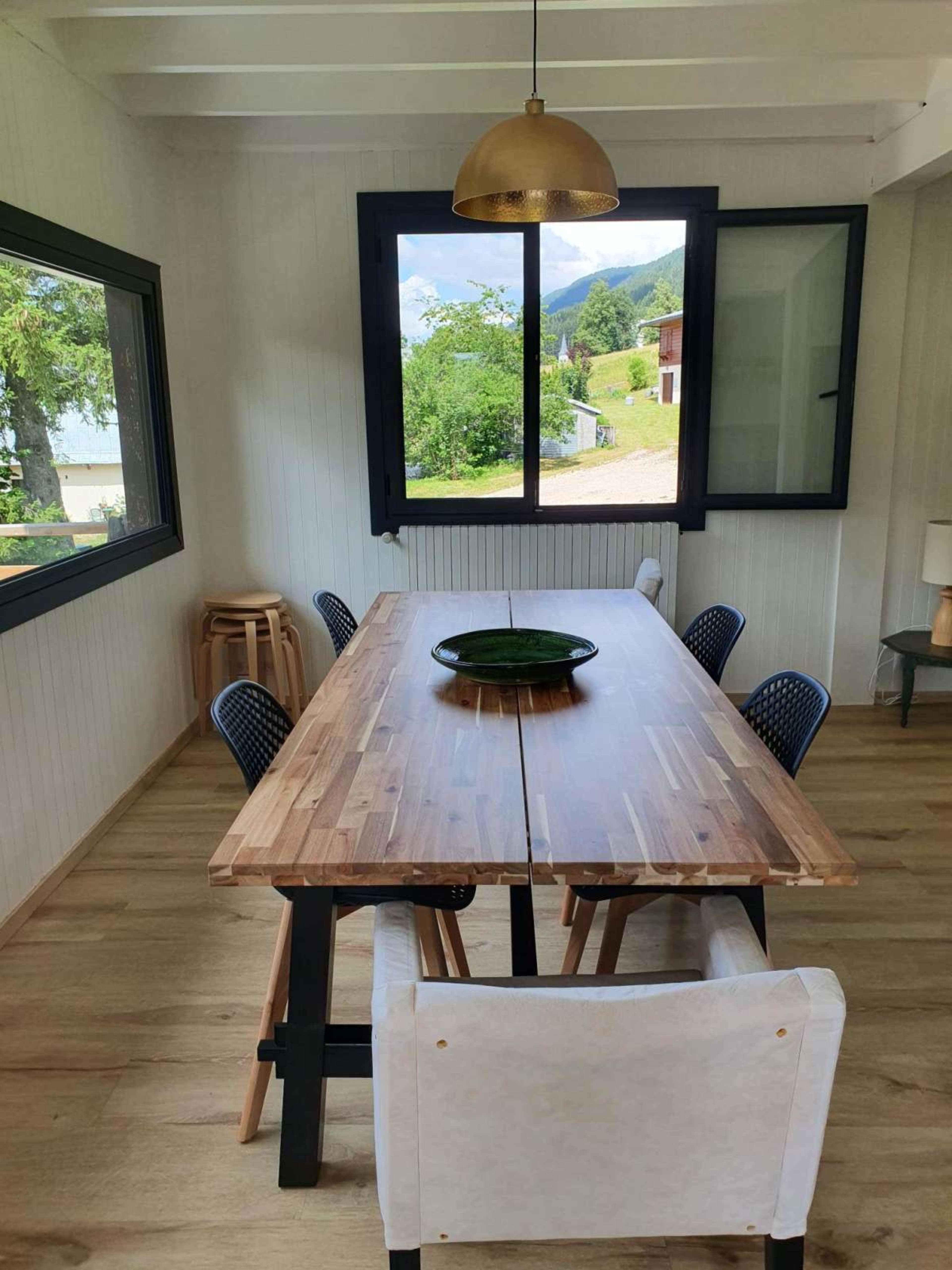 The image shows a dining area with a wooden table surrounded by chairs, large windows providing natural light, and a pendant light hanging above.