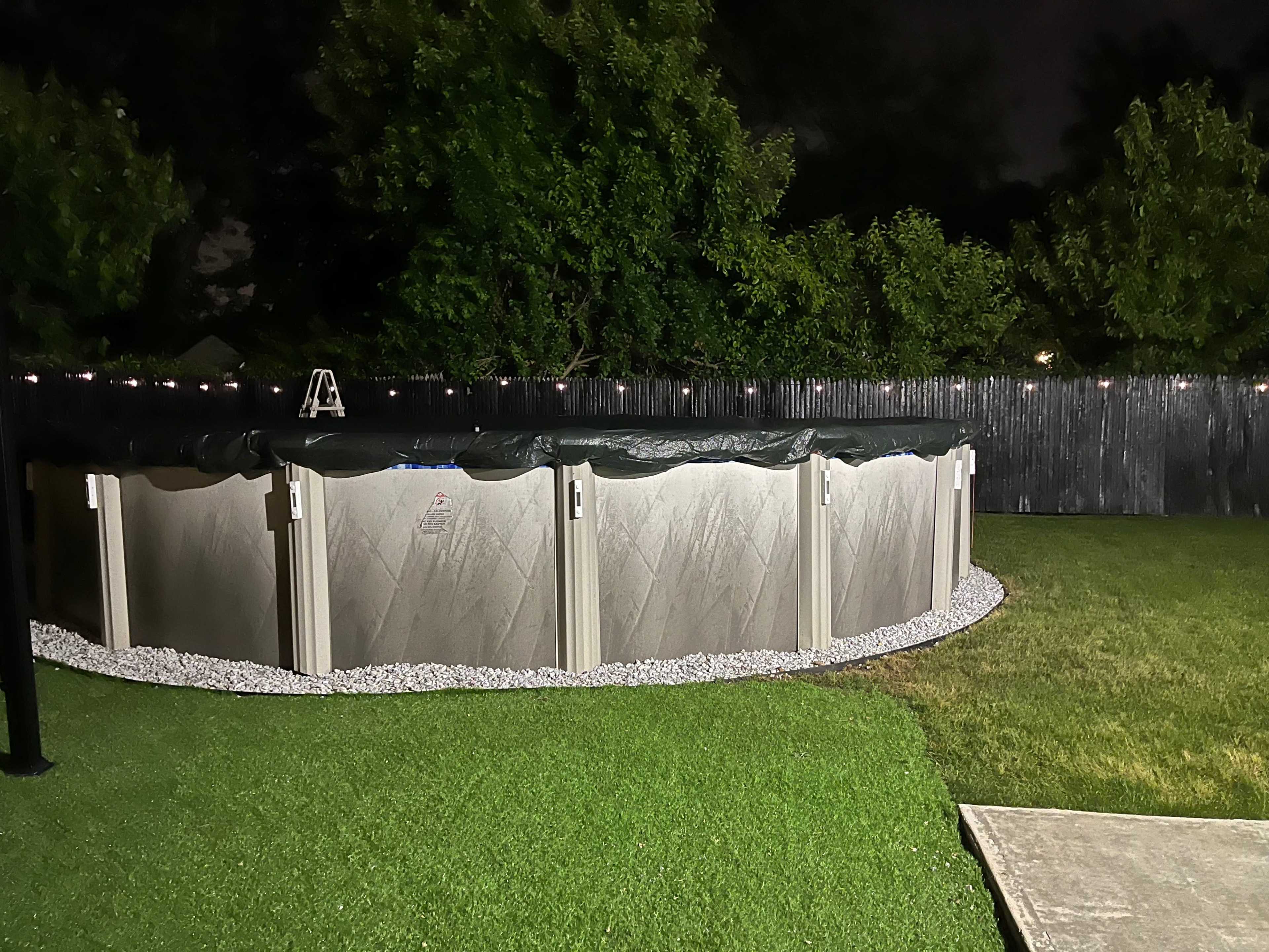 A round above-ground pool is surrounded by gravel and sits in a backyard at night, with a wooden fence and trees in the background.