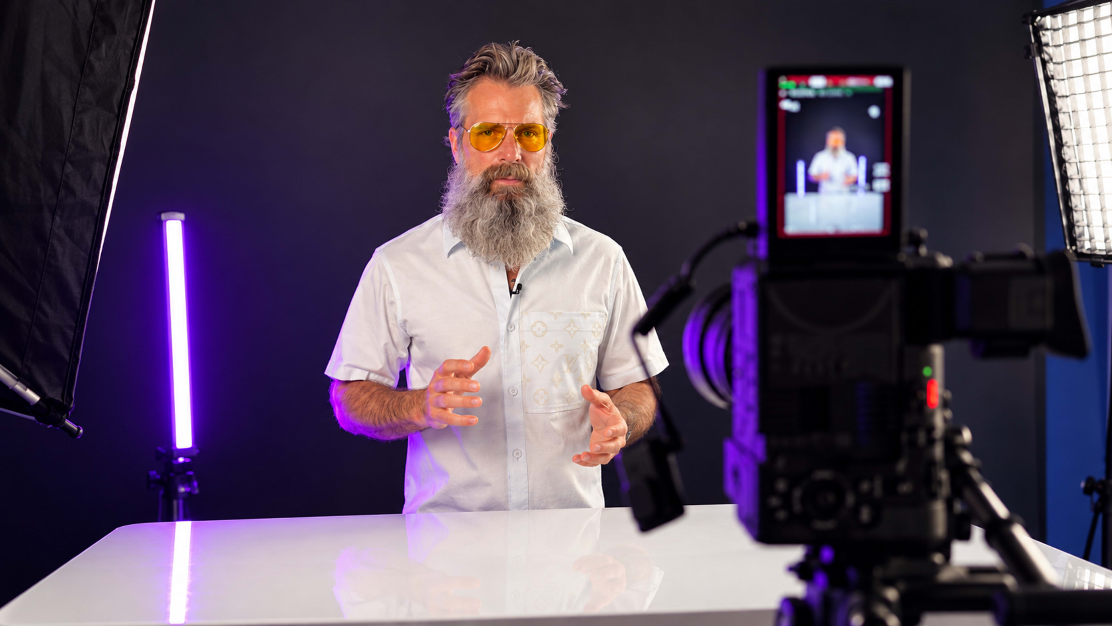 A man with a beard and yellow sunglasses gestures while standing behind a white table in front of a camera on a tripod.