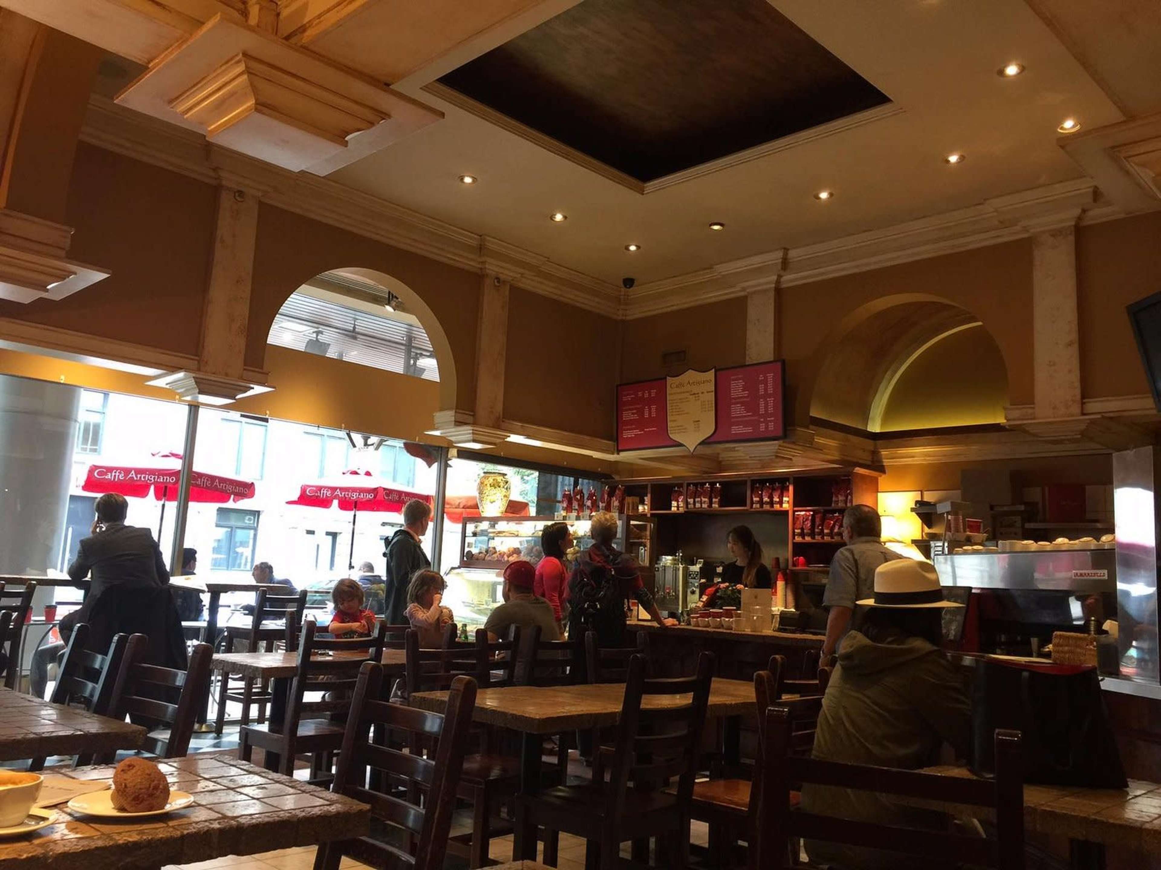 A bustling café interior features customers seated at wooden tables, with staff at the counter preparing orders behind a glass display.