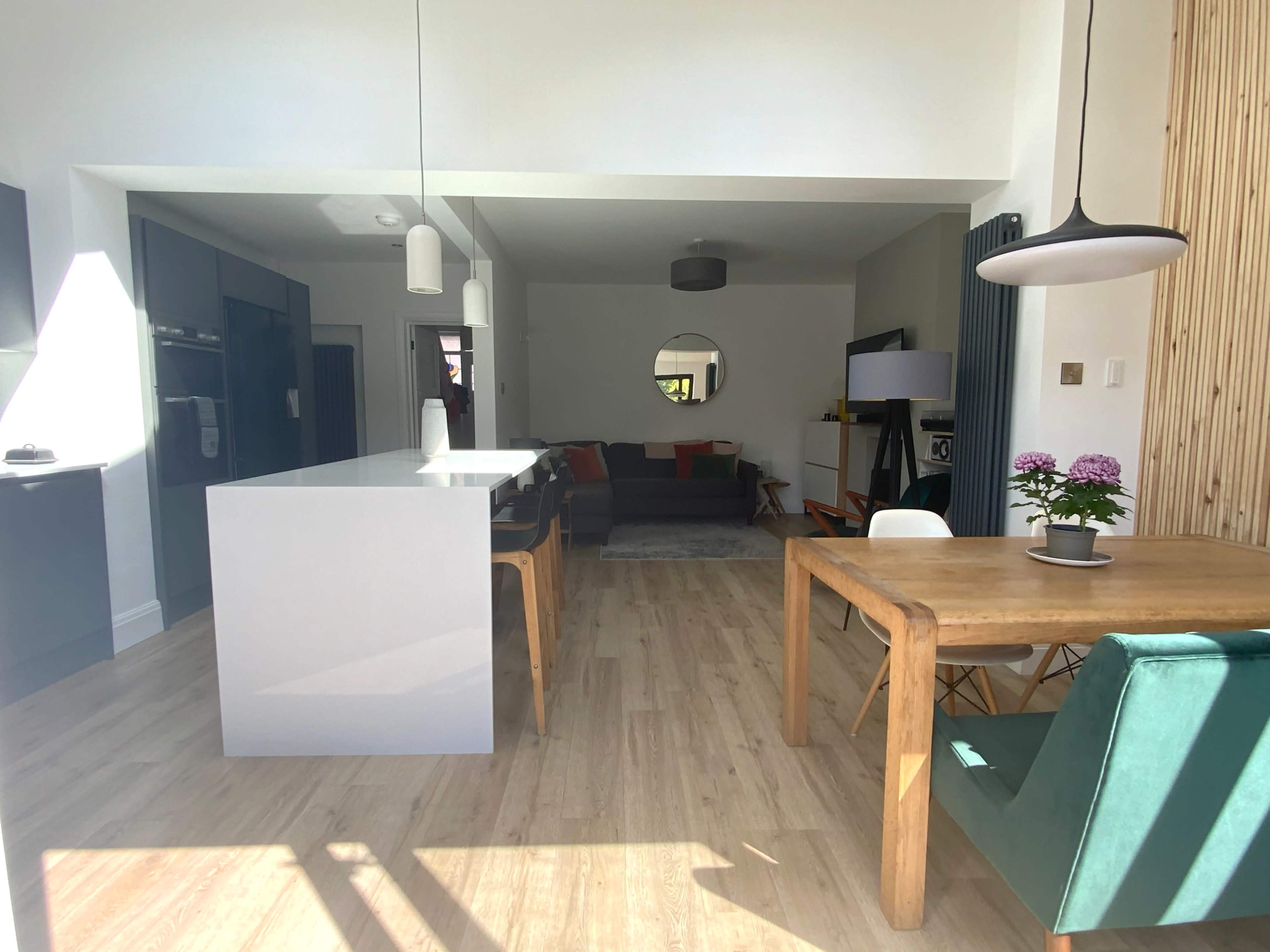 A modern open-concept kitchen and living area featuring a light wood dining table, a gray sofa, and a bright window that allows sunlight to illuminate the space.