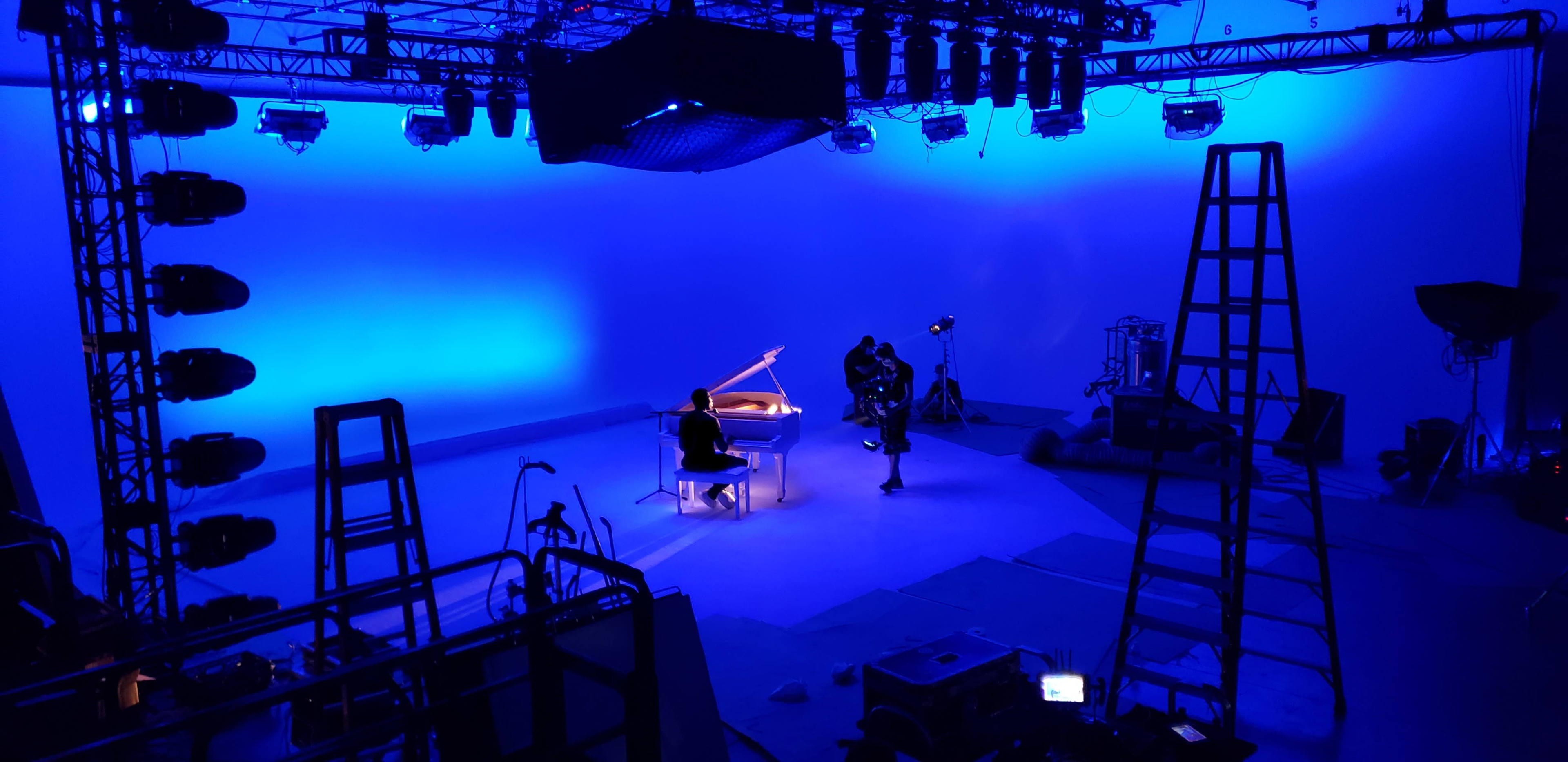 A music studio is set up with a grand piano in the center, illuminated by blue lighting, surrounded by cameras and various equipment.