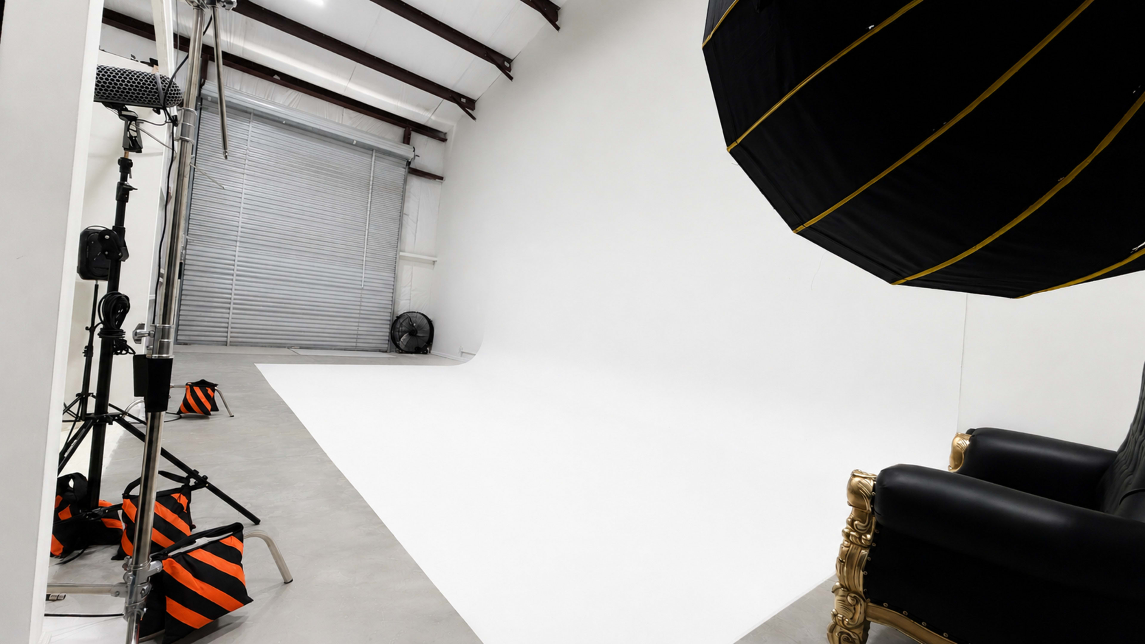 The image shows a photography studio featuring a white backdrop, a large softbox light, and a decorative black chair with gold accents.
