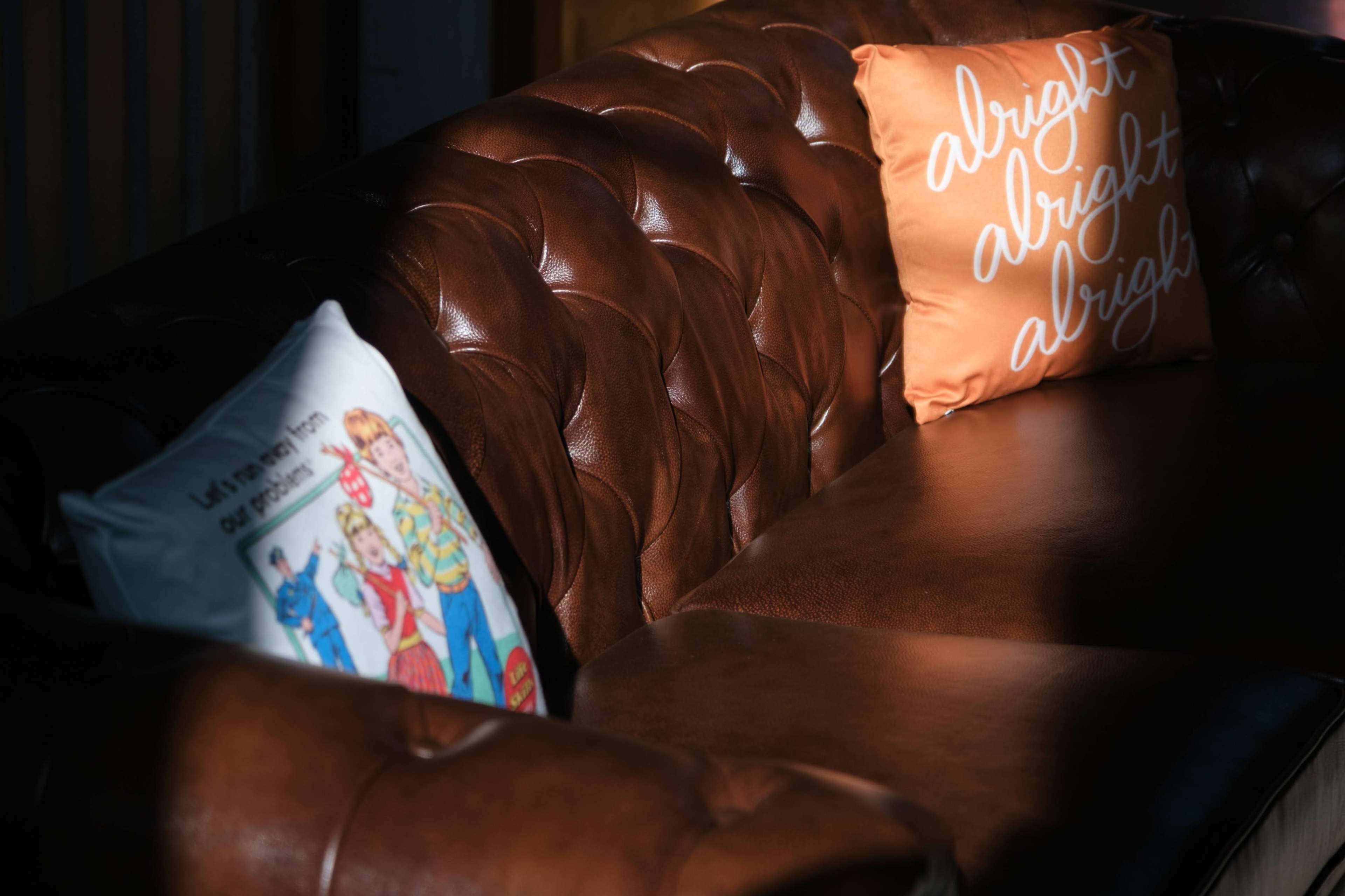 A brown leather couch with tufted upholstery is adorned with two decorative pillows, one featuring a colorful illustration and the other with the word "alright" in script.