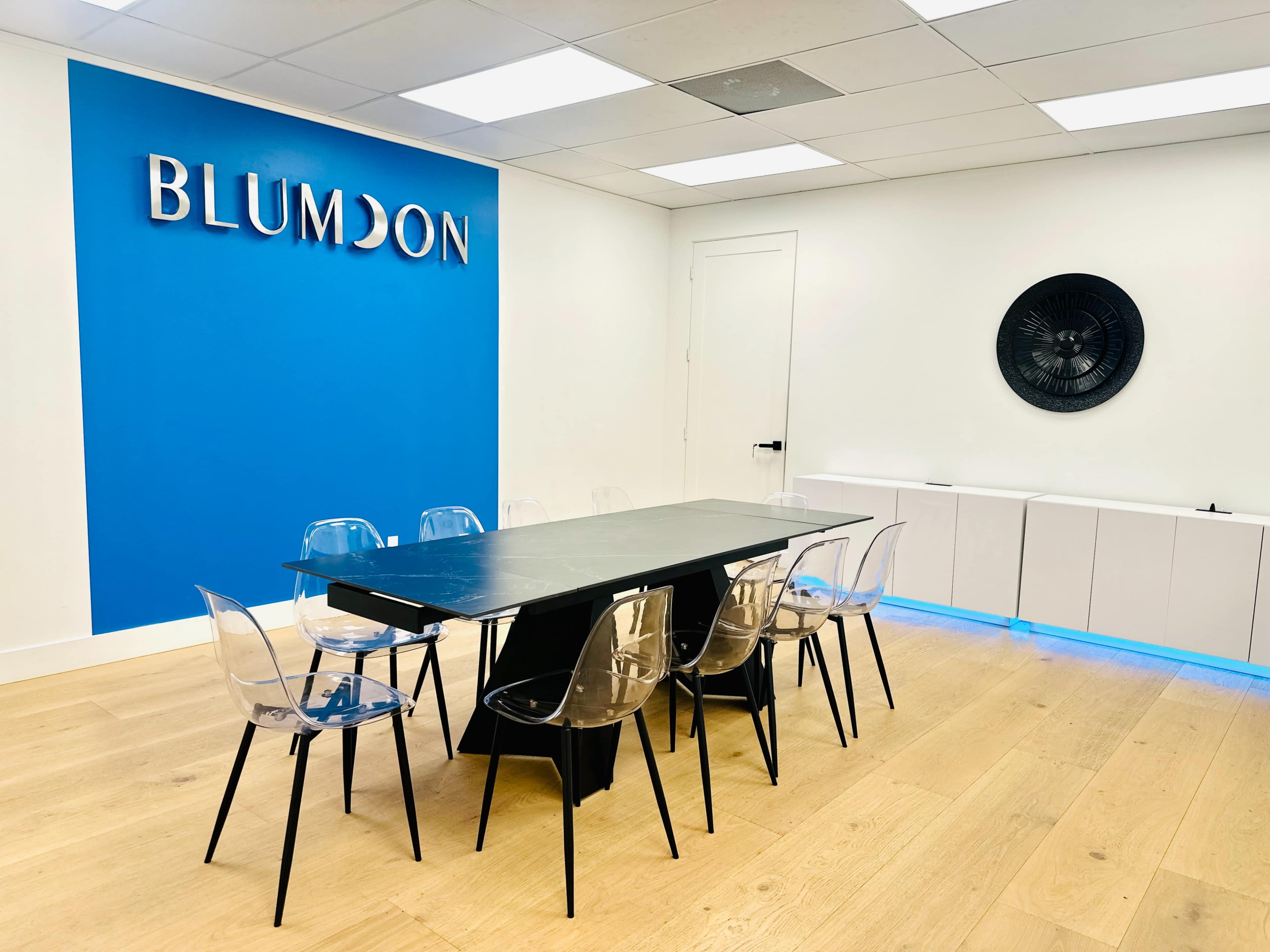 A modern conference room features a long black table surrounded by clear plastic chairs, with a blue accent wall displaying the name "BLUMDON."