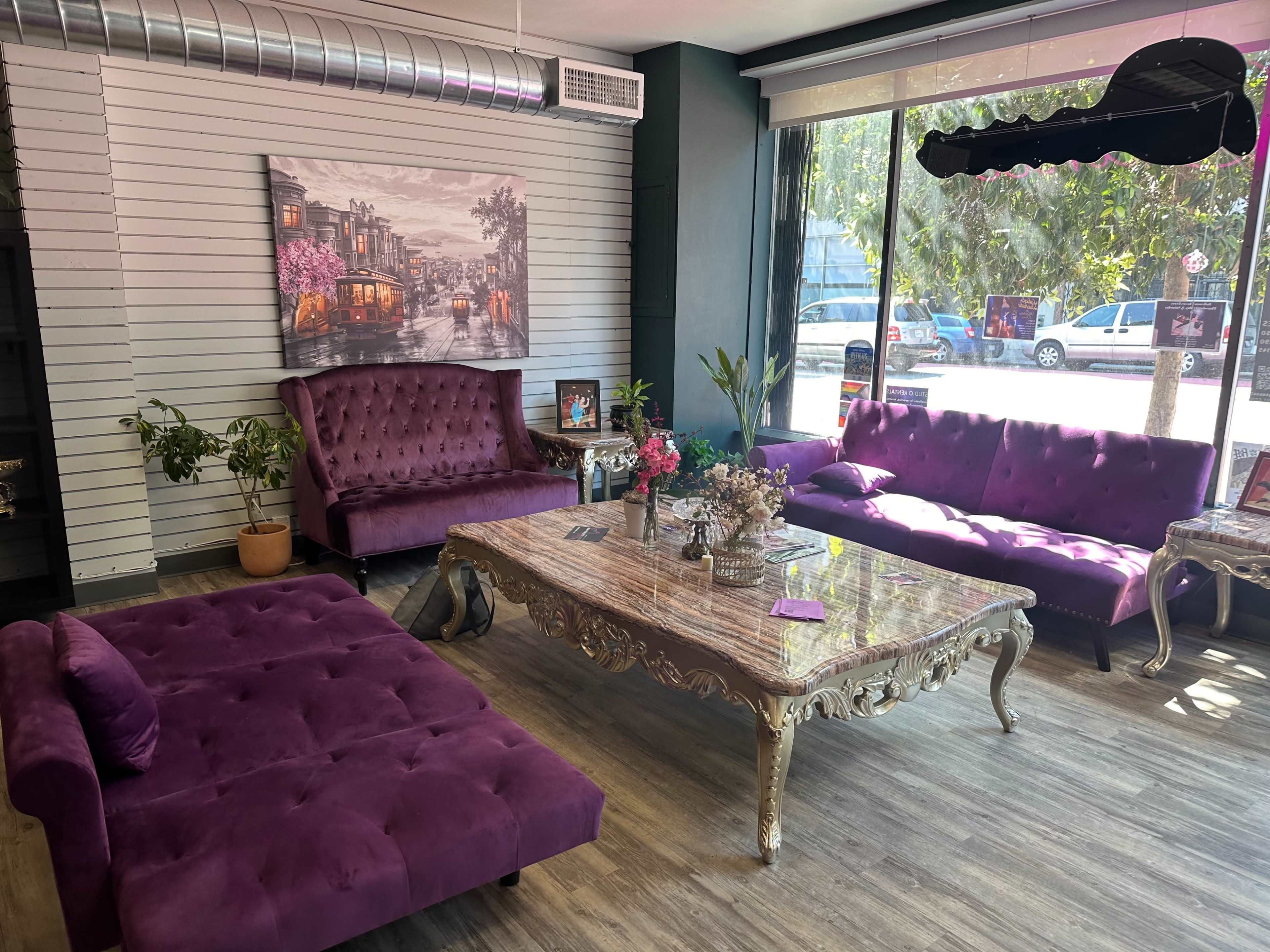 A stylish waiting area featuring purple velvet furniture, including sofas and a coffee table, beside large windows with natural light.