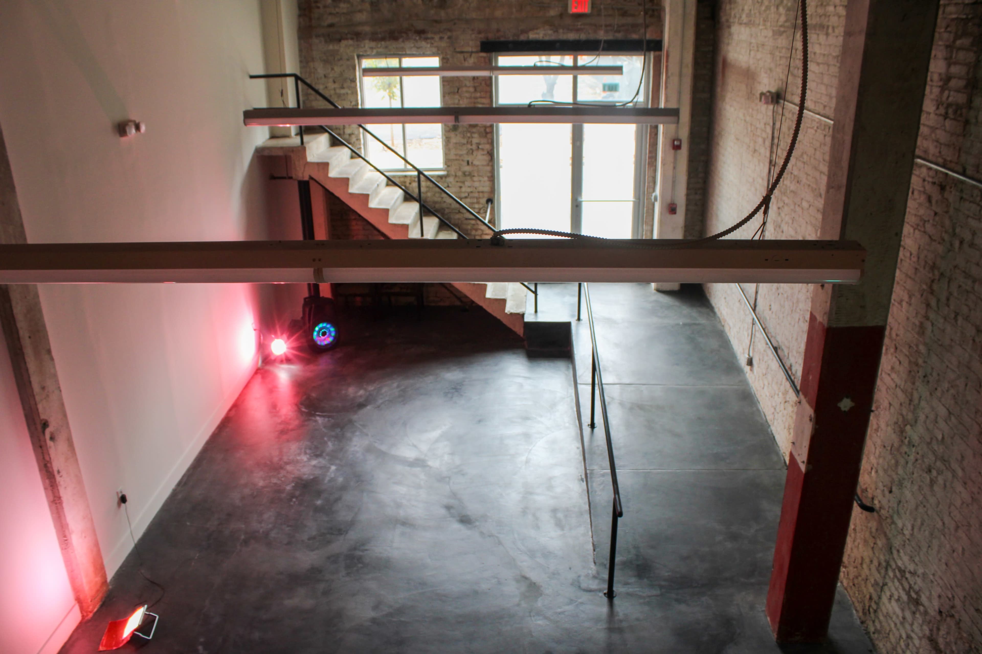 The image shows a spacious, empty interior space featuring exposed brick walls, a staircase, and lighting elements.