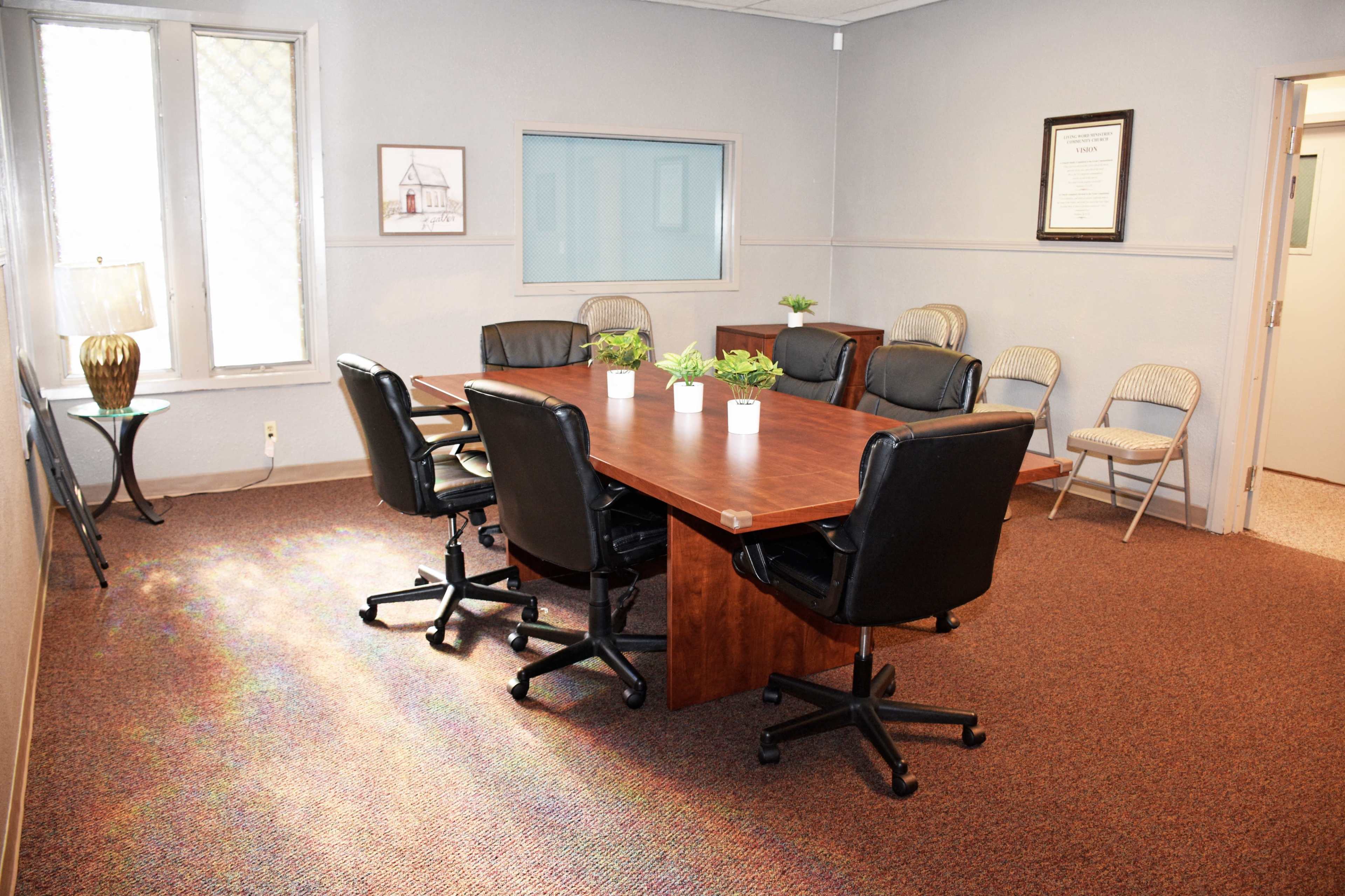 A conference room features a large wooden table surrounded by black rolling chairs, with additional folding chairs positioned against the wall.
