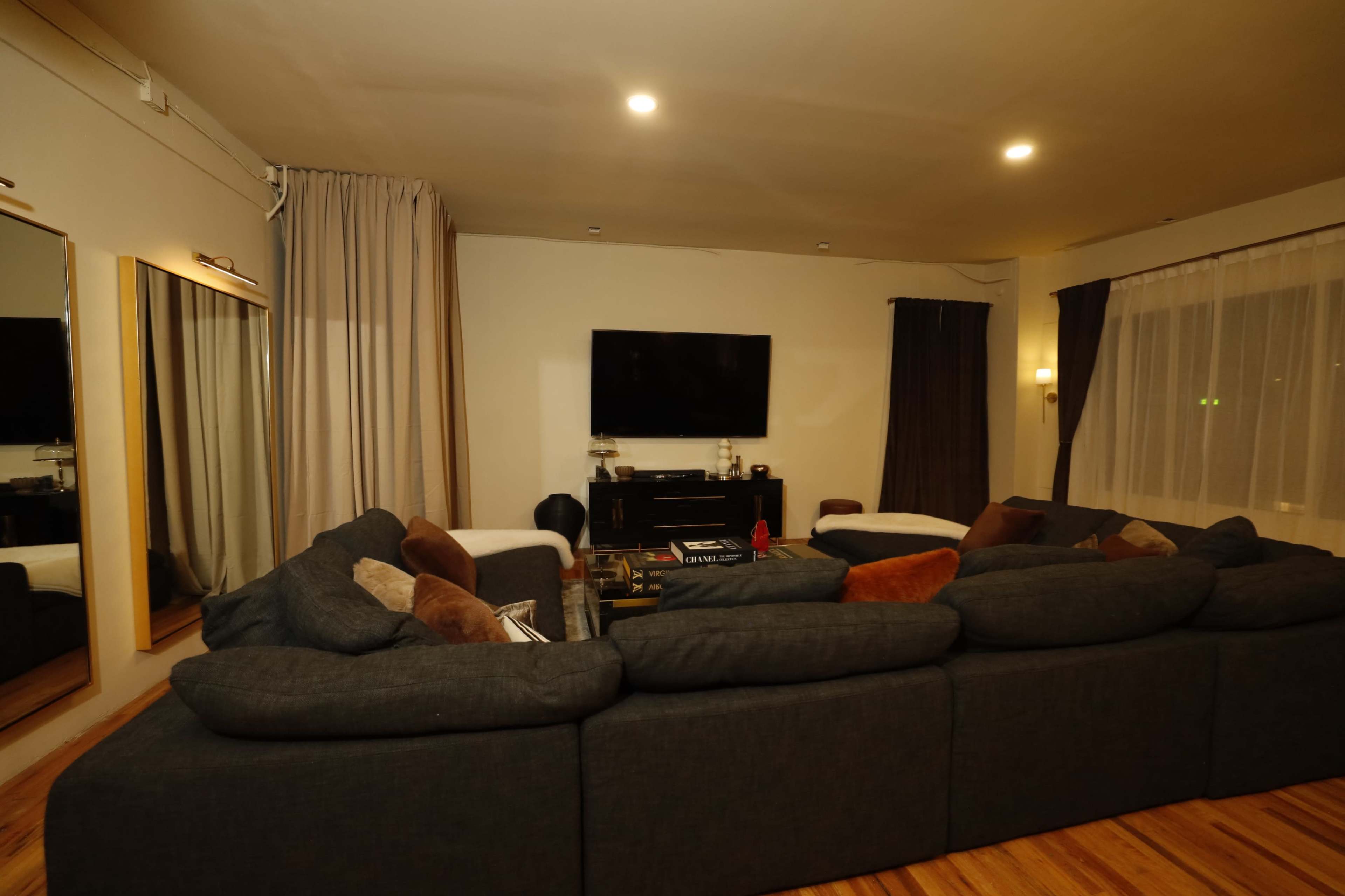The image shows a living room with a large sectional sofa, a dark television mounted on the wall, and curtains on the windows.