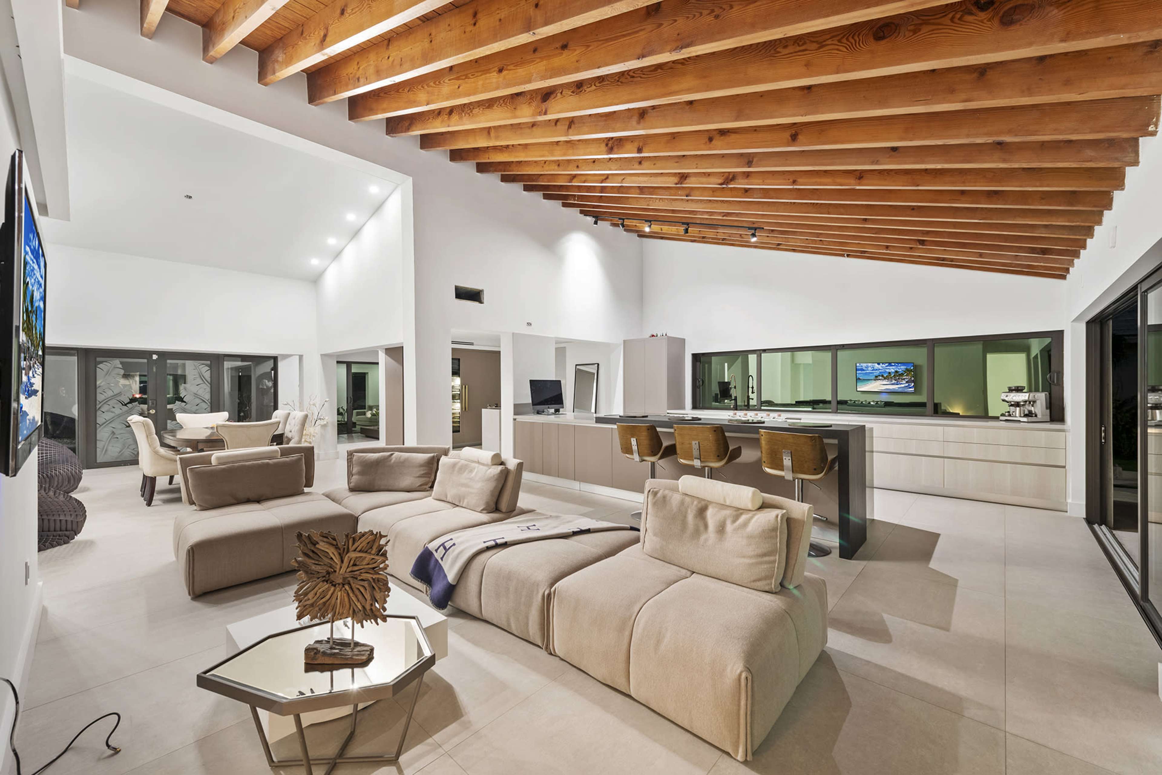 A modern open-concept living space with a kitchen, dining area, and lounge, highlighted by wooden beams and large windows.