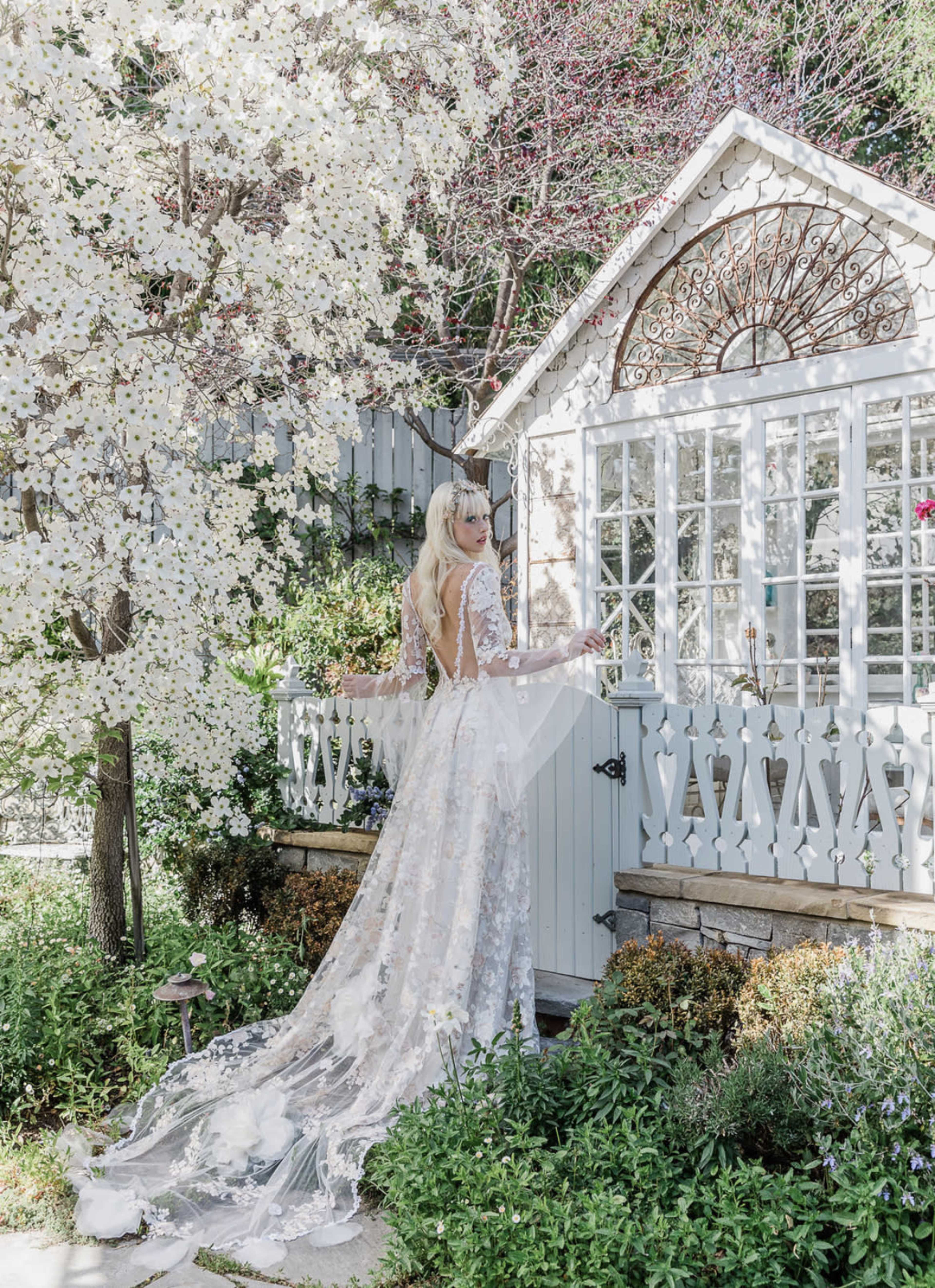 A woman in a detailed lace wedding dress stands by a vintage-style garden shed, surrounded by blooming white flowers and greenery.