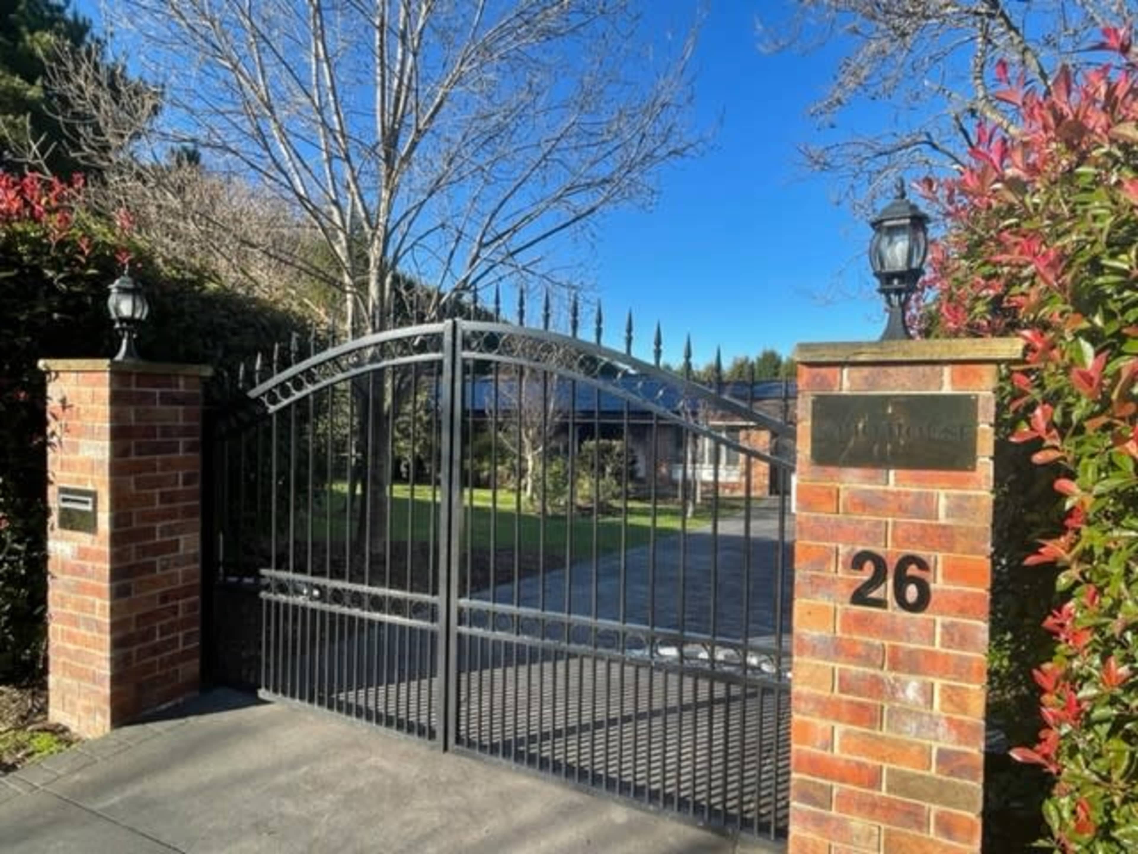 A black wrought iron gate with stone pillars stands at the entrance of a driveway marked with the number 26, leading to a landscaped property.
