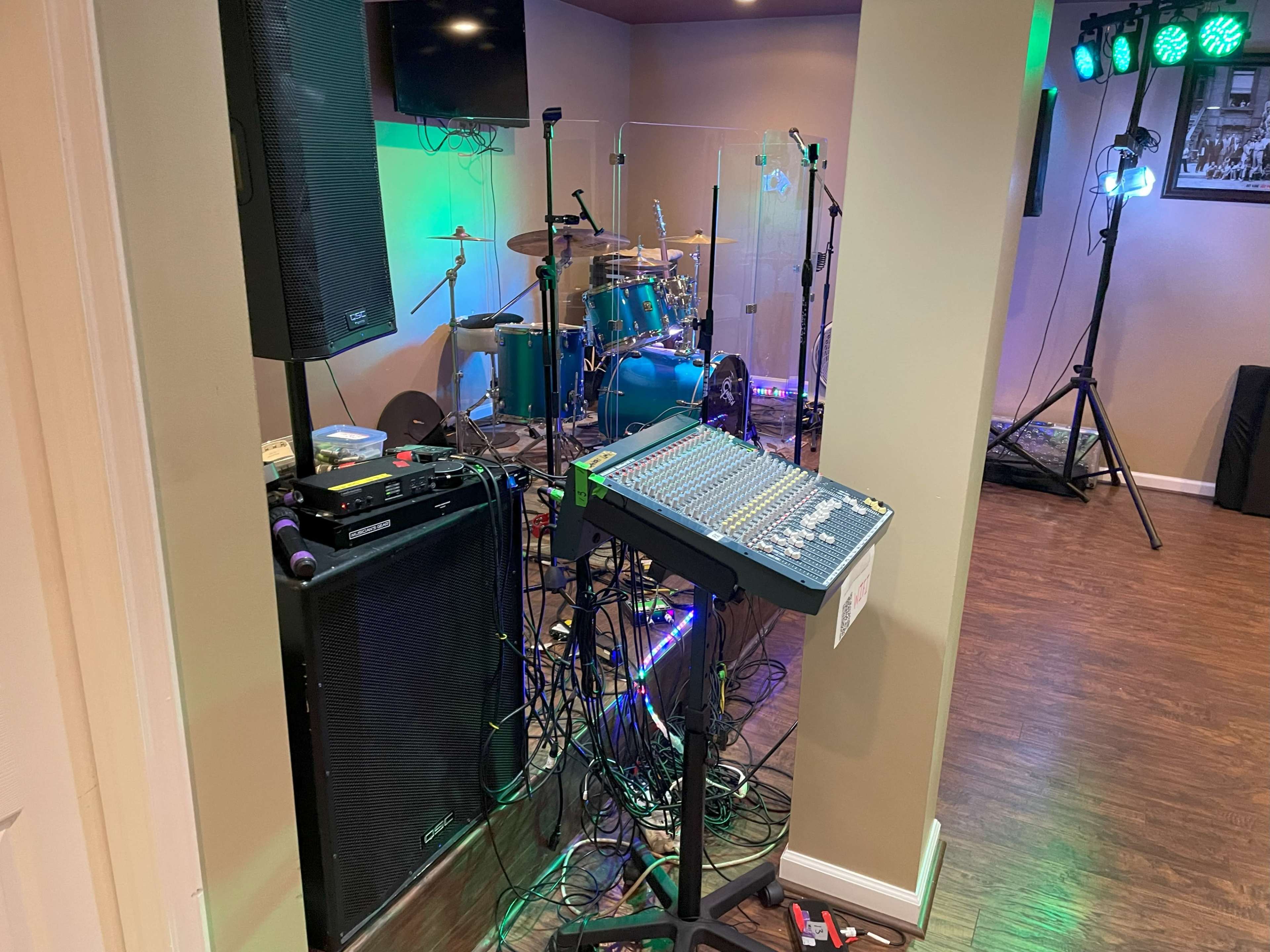 A music setup features a mixing console and a speaker next to a drum kit and stage lights within a room.