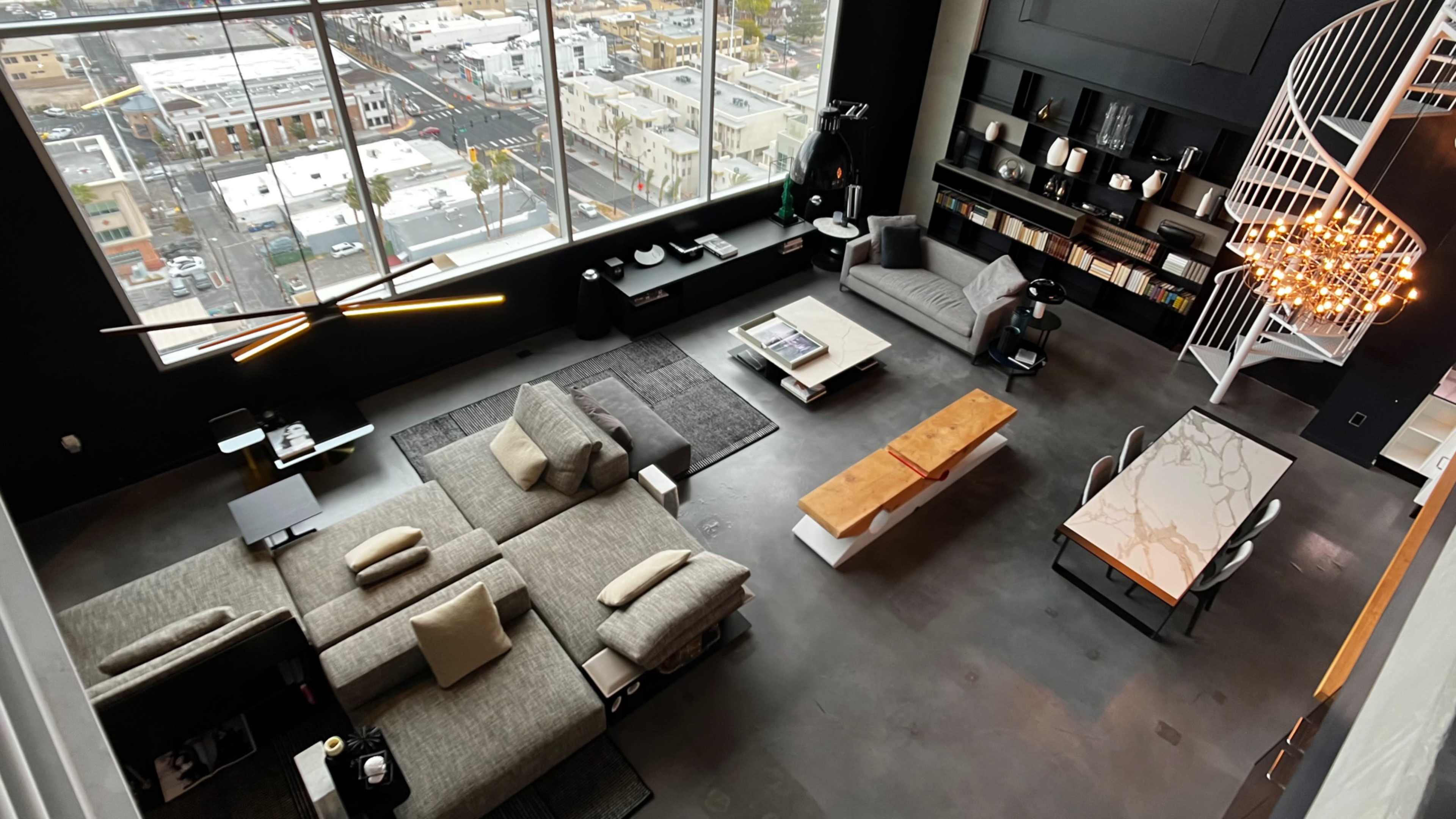 A modern living space with large windows, featuring a grey sectional sofa, a white dining table with a marble top, and a spiral staircase.