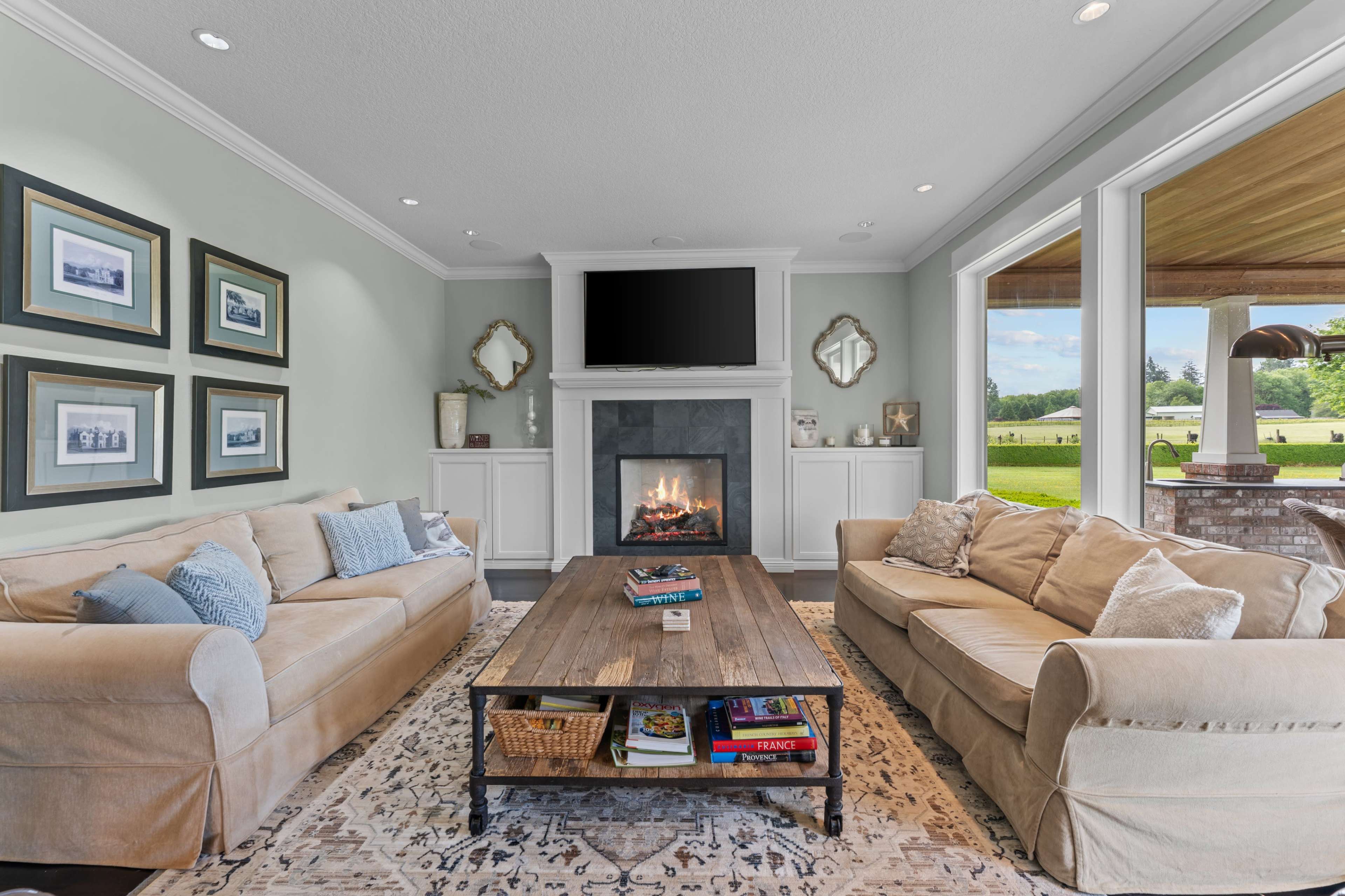 The living room features two beige sofas, a wooden coffee table, a stone fireplace with a mounted television above, and large windows that provide a view of the outdoors.